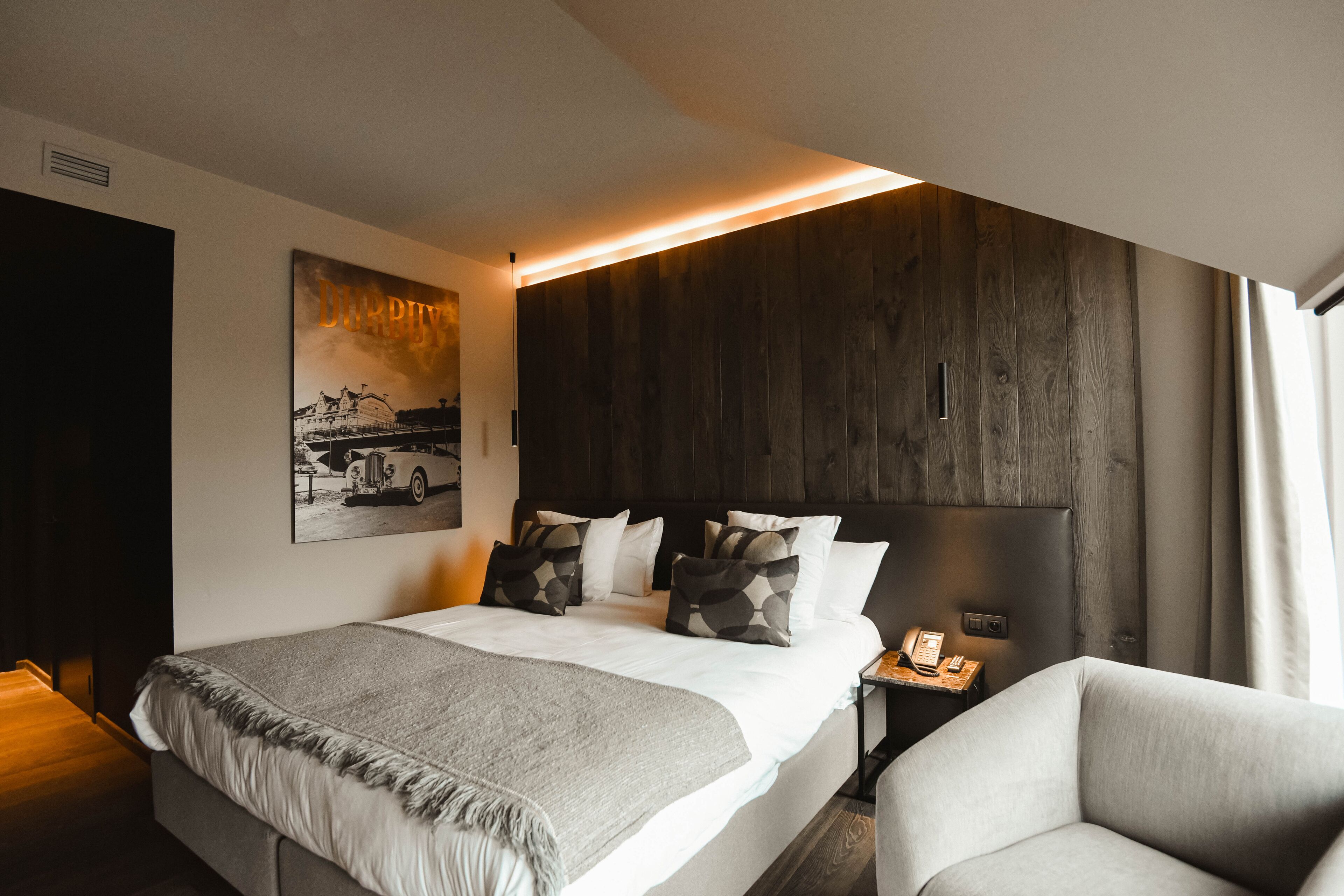A modern hotel room with a large bed, a bedside table with a lamp, a chair, a dark wood headboard, and a framed picture on the wall.