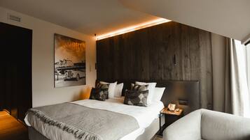 A modern hotel room with a large bed, a bedside table with a lamp, a chair, a dark wood headboard, and a framed picture on the wall.