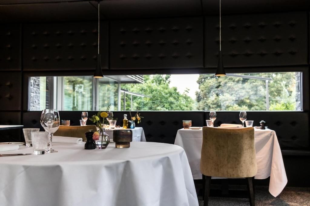 Un restaurante con mesas redondas, manteles blancos y vistas al exterior con vegetación.
