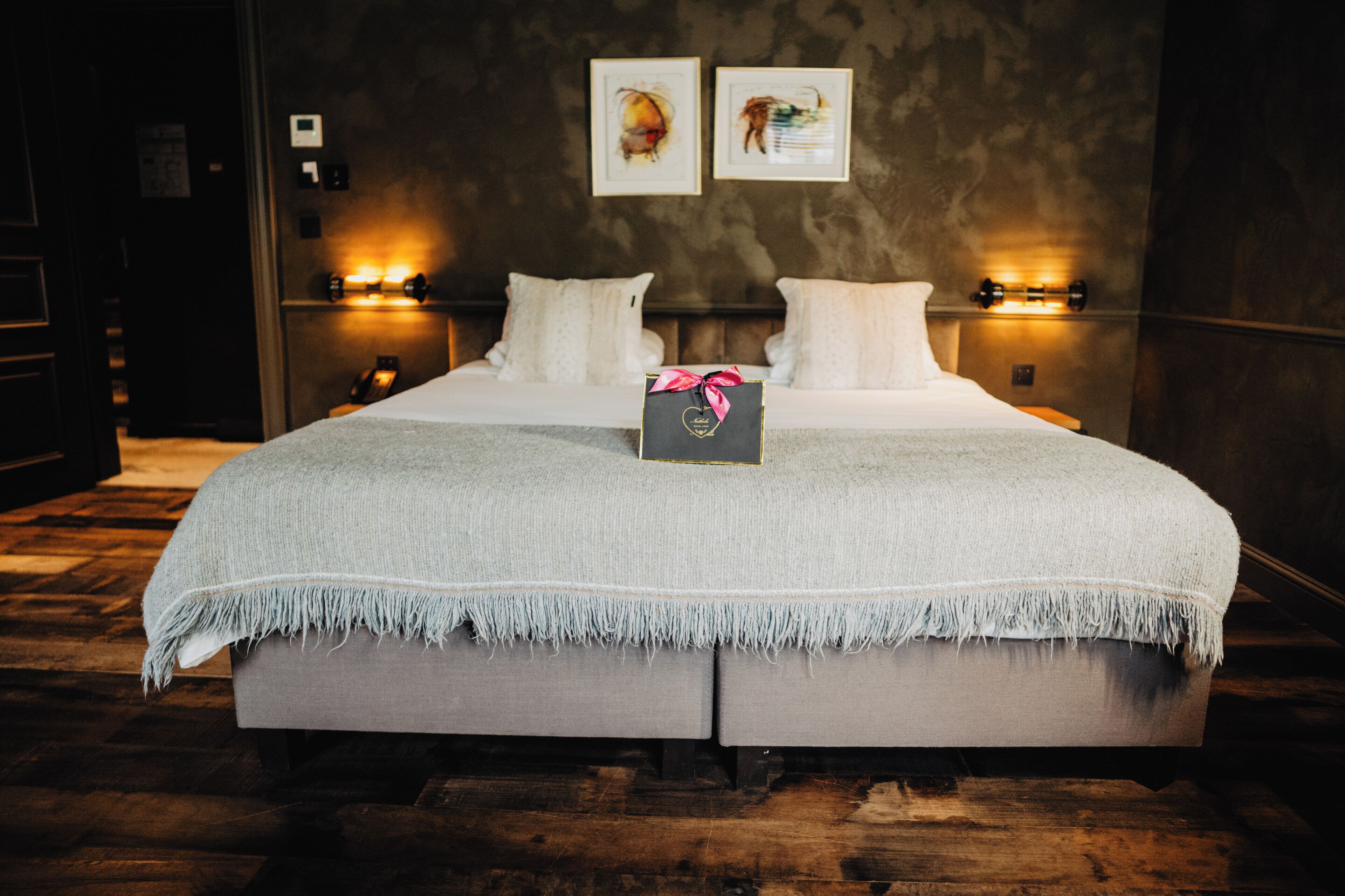Una cama doble con una manta gris, dos lámparas de noche y una caja de regalo sobre la cama.