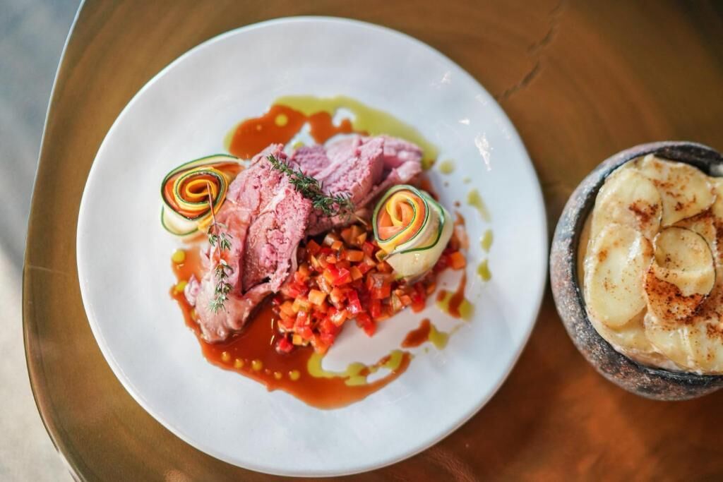 Un plato de comida con carne cortada, verduras y un recipiente pequeño que parece ser de puré de patatas.