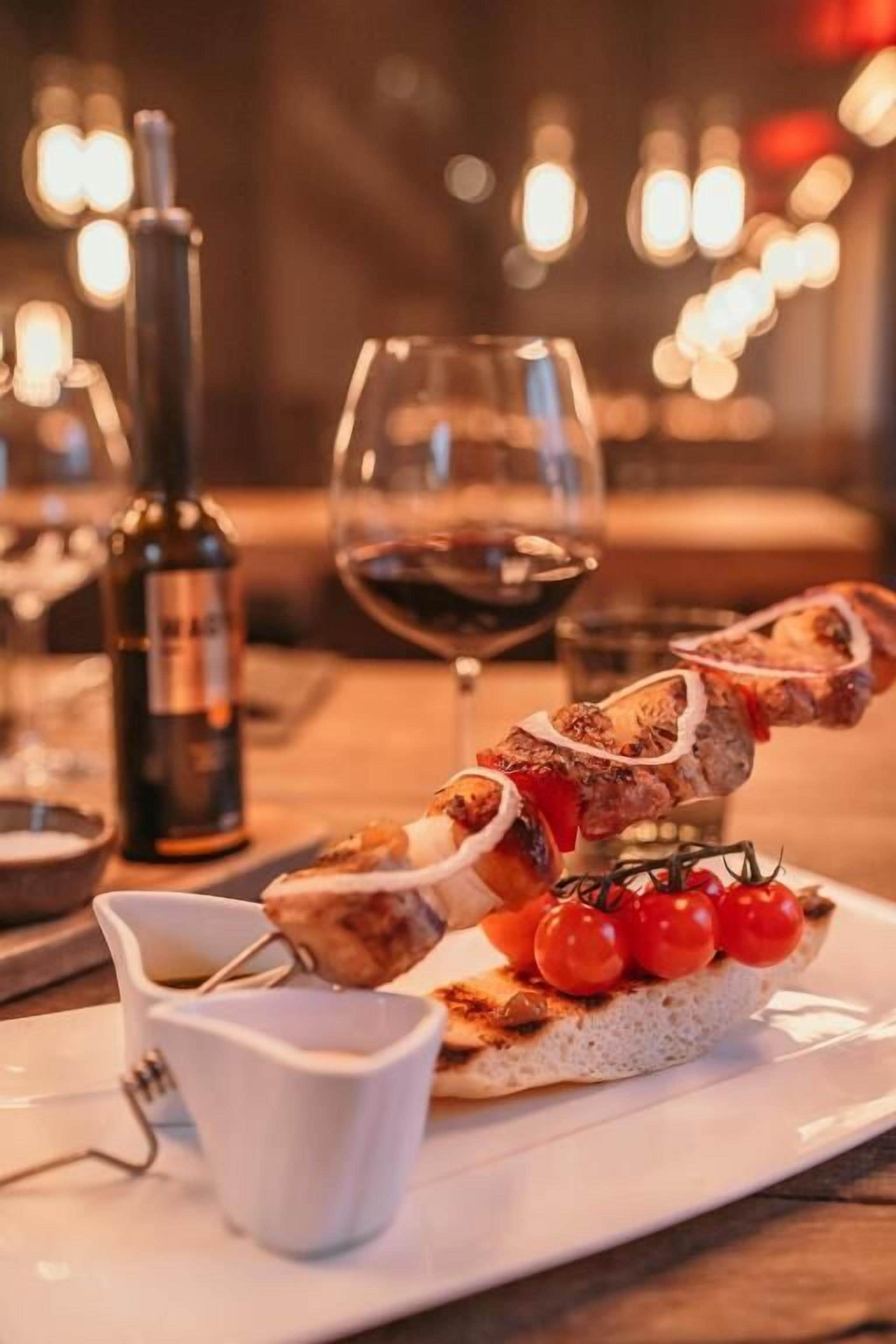 A plate with skewered meat, cherry tomatoes, and a small bowl of sauce, accompanied by a bottle of wine and a glass.