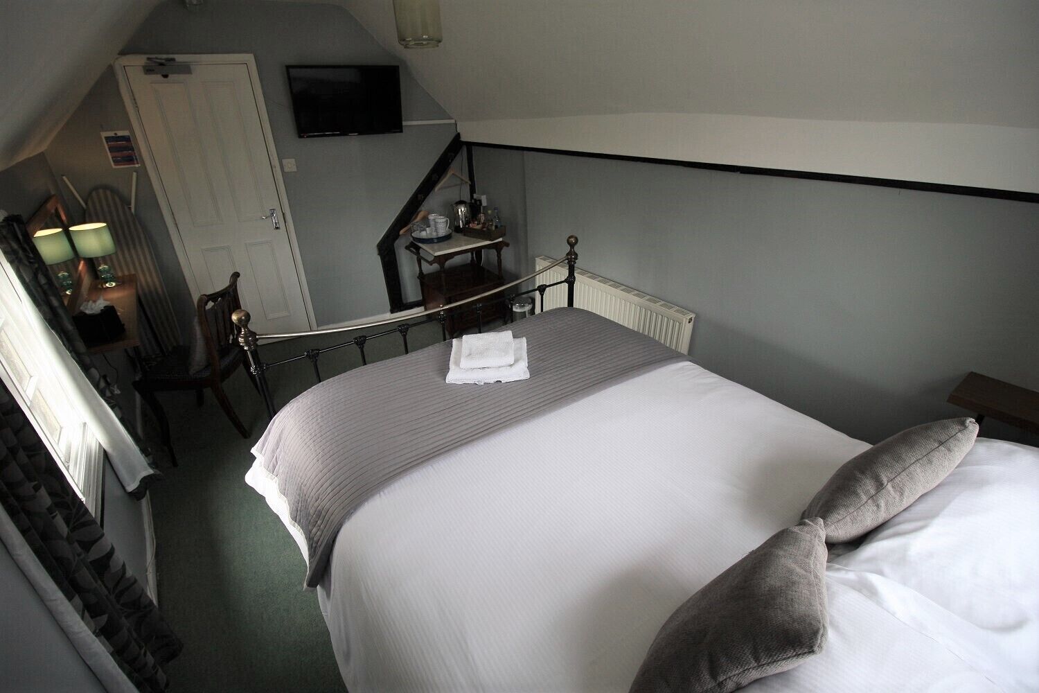Een net opgemaakt bed met een wit-grijs gestreept dekbedovertrek, een grijs kussen en een opgevouwen witte handdoek. Tegen de muur staat een klein tafeltje met een lamp en een dienblad met spullen.