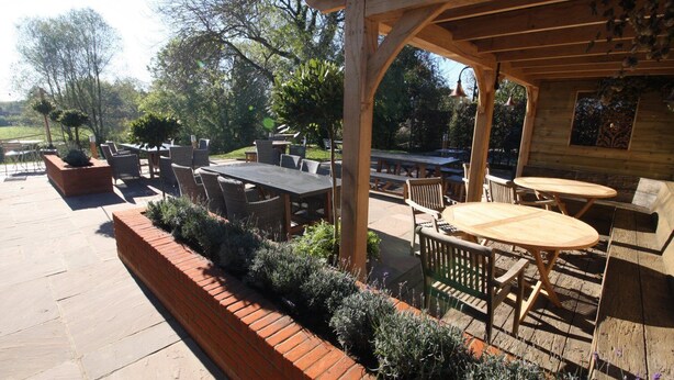 An outdoor patio with wooden tables and chairs, a brick planter with shrubs, and a pergola structure. - The Carpenters Arms (Newbury)