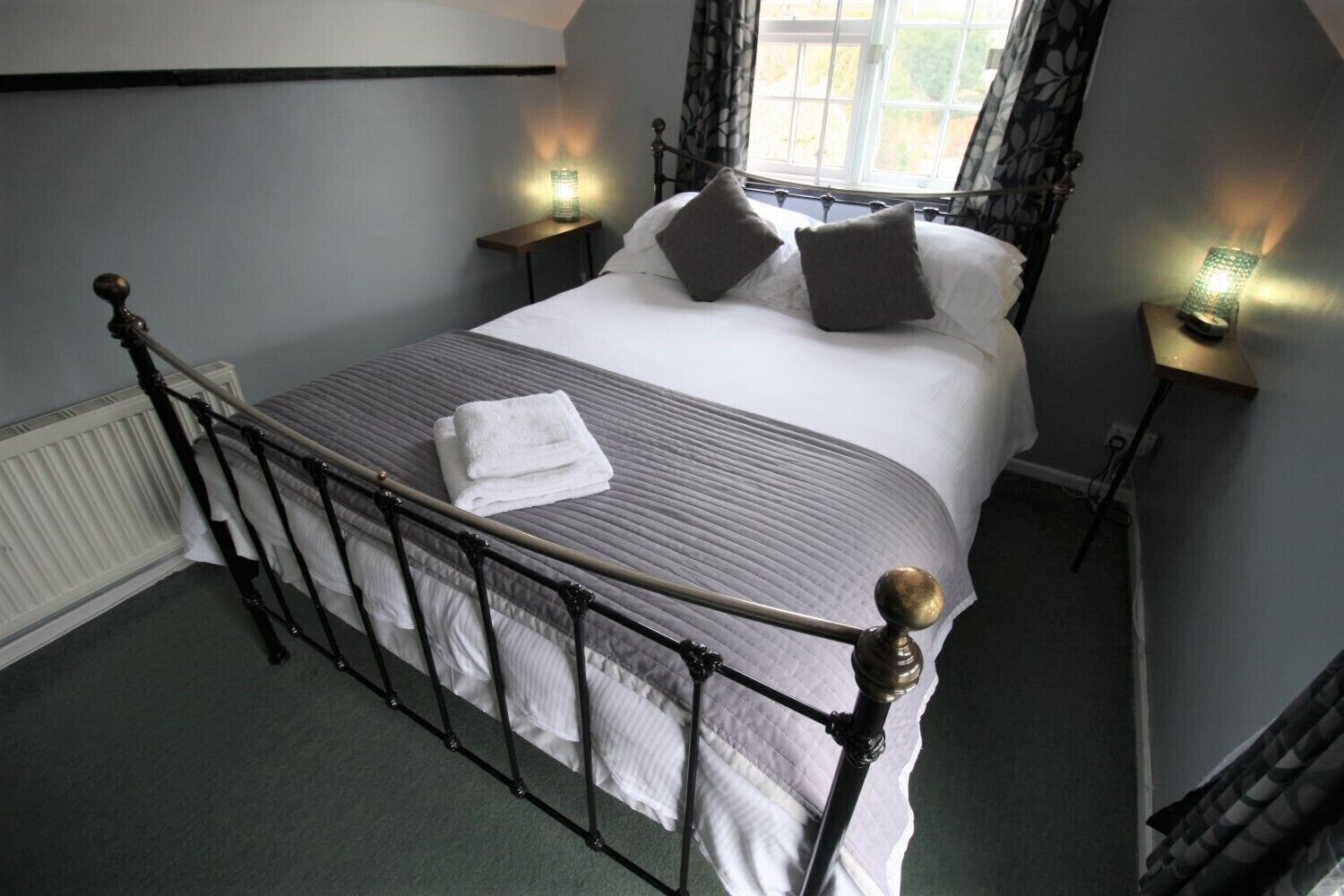 A neatly made bed with a white comforter and pillows, a folded white towel, and a metal headboard. There are two bedside tables with lamps, one on each side of the bed.