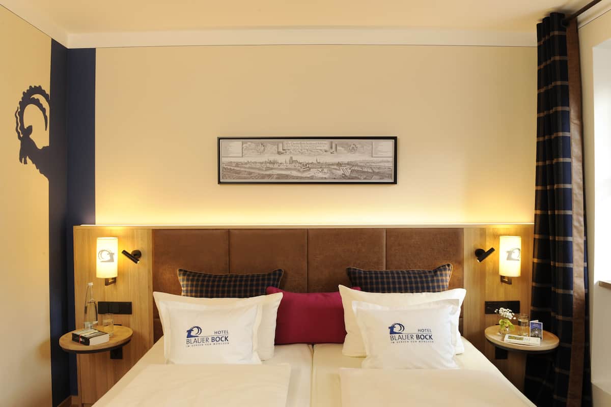 A hotel room with a double bed, two pillows with the hotel logo, a framed cityscape picture, and two bedside tables with lamps.