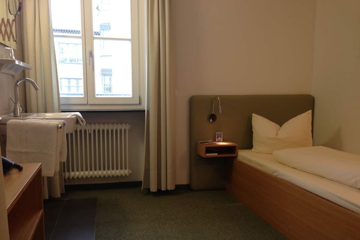 A hotel room with a bed, a bedside table with a lamp, a radiator, a window with curtains, and a wooden dresser.