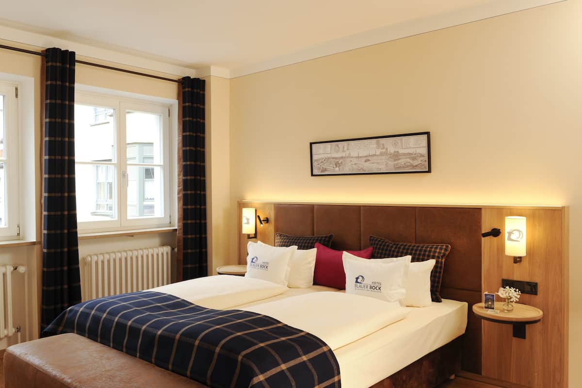 A hotel room with a large bed, two bedside tables, a headboard, a framed picture on the wall, and a window with curtains.