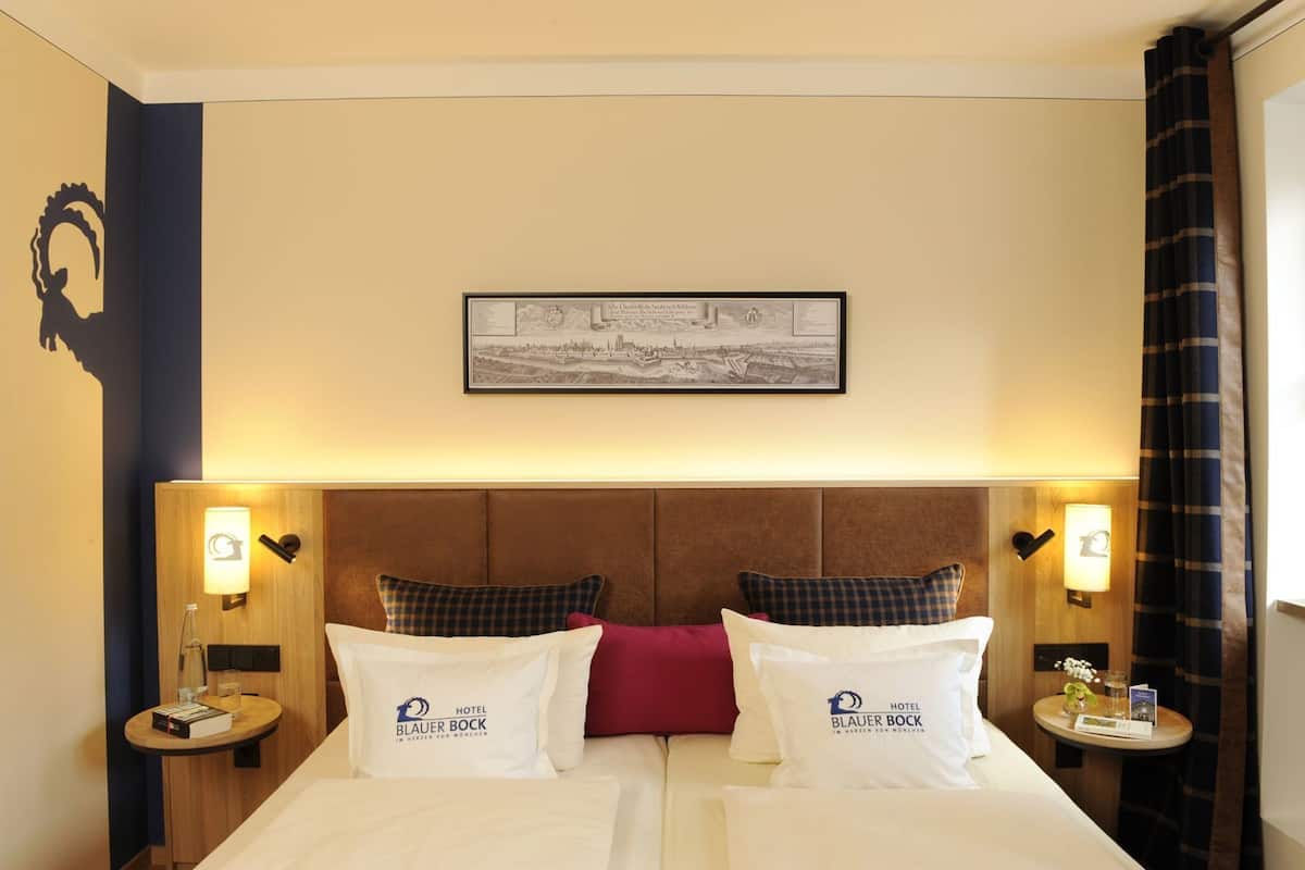 A hotel room with a double bed, two pillows with the hotel's logo, a framed cityscape picture, and two bedside tables with lamps.