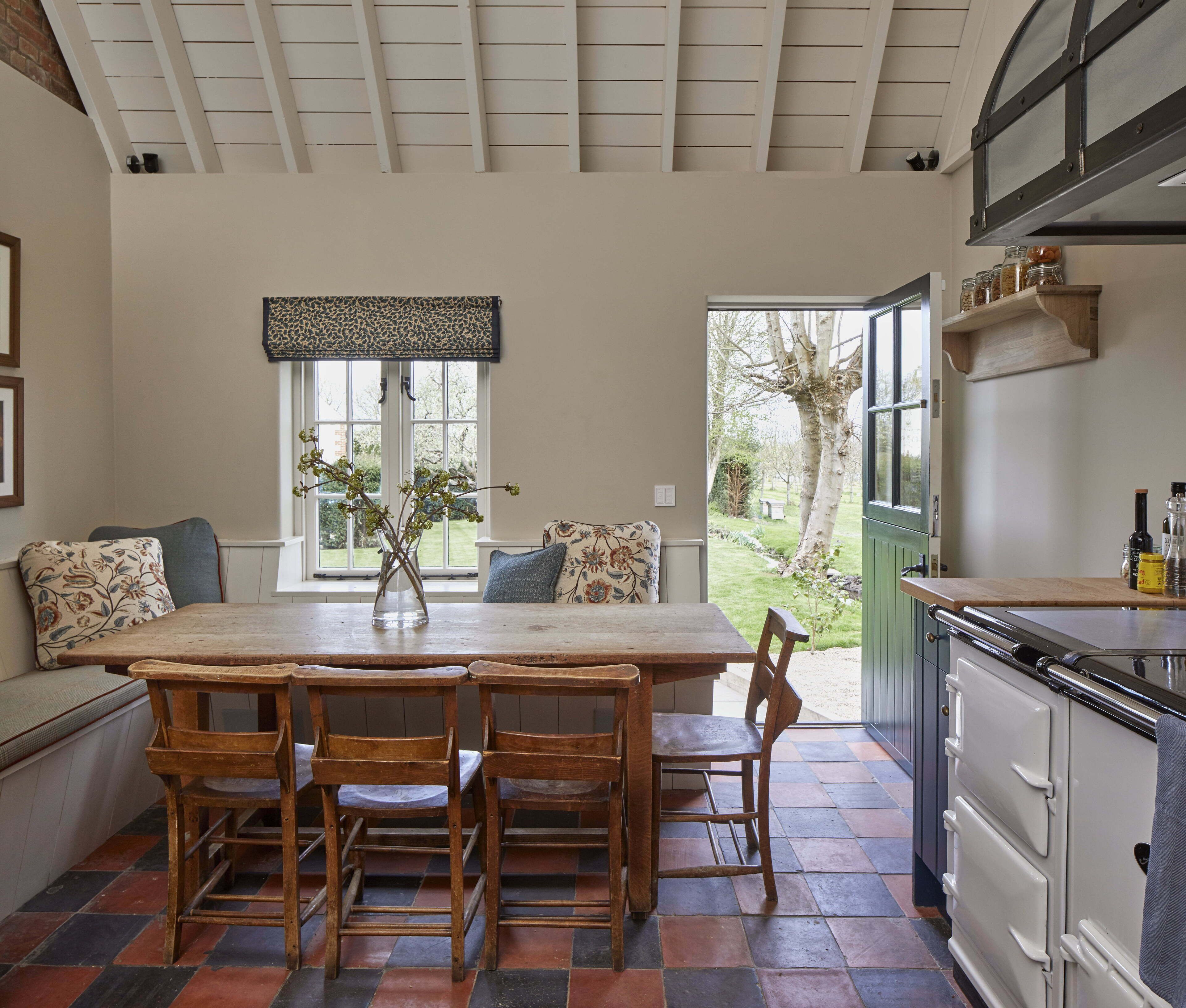 A dining area with a wooden table and chairs, a vase with flowers, and a view of the garden through a window.
