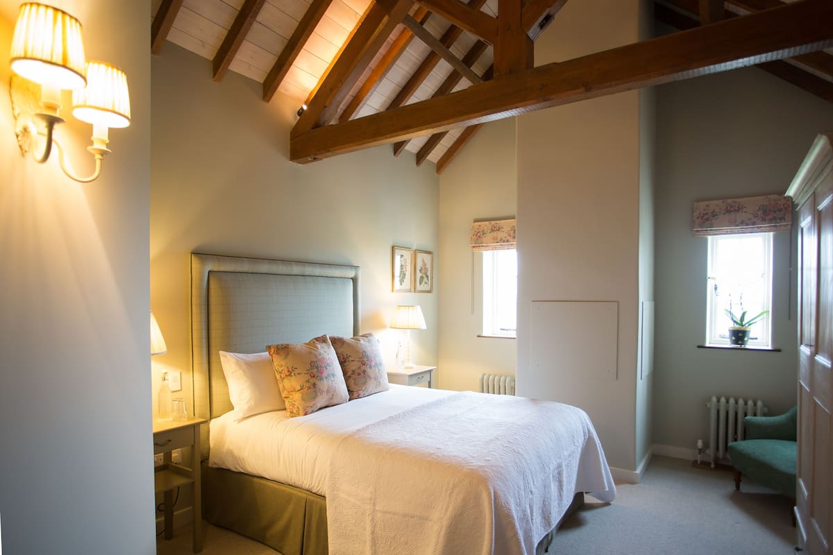 A bedroom with a large bed, wooden beams, a nightstand, a lamp, a window with a plant, and a radiator.