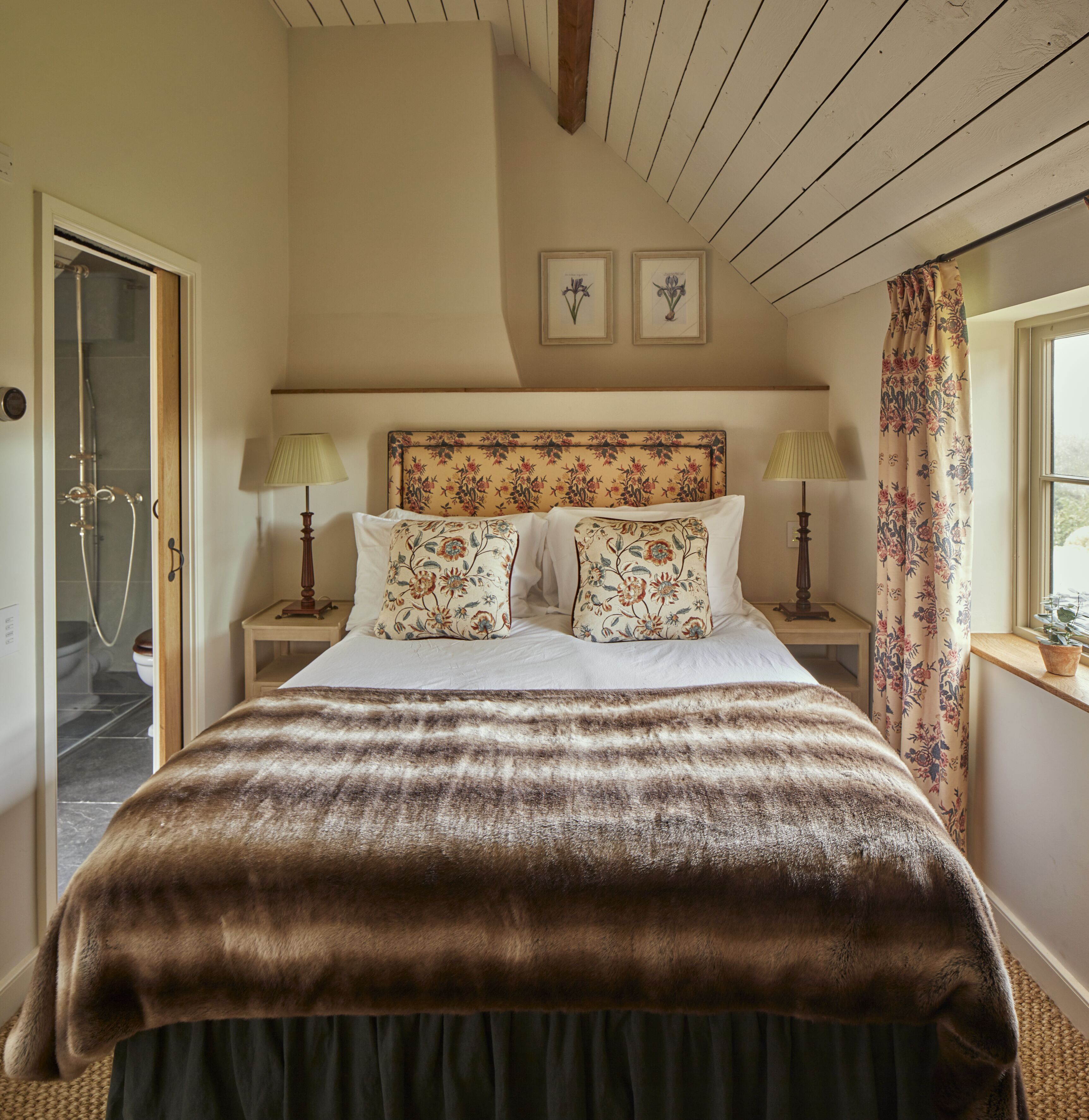 A bedroom with a large bed, floral headboard, two bedside lamps, and a window with curtains.