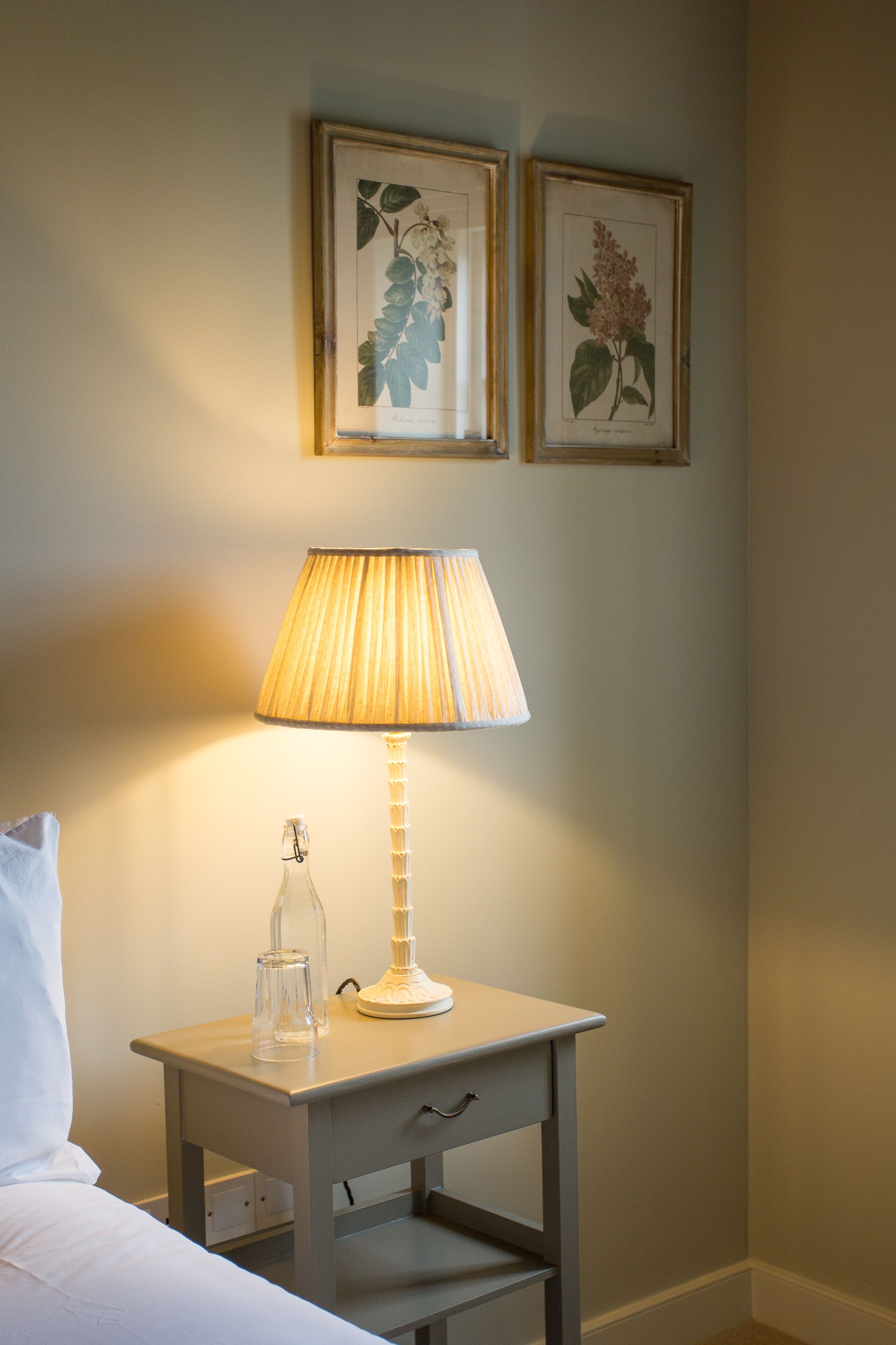A neatly made bed with a white pillow, a bedside table with a lamp, and two framed botanical prints on the wall.