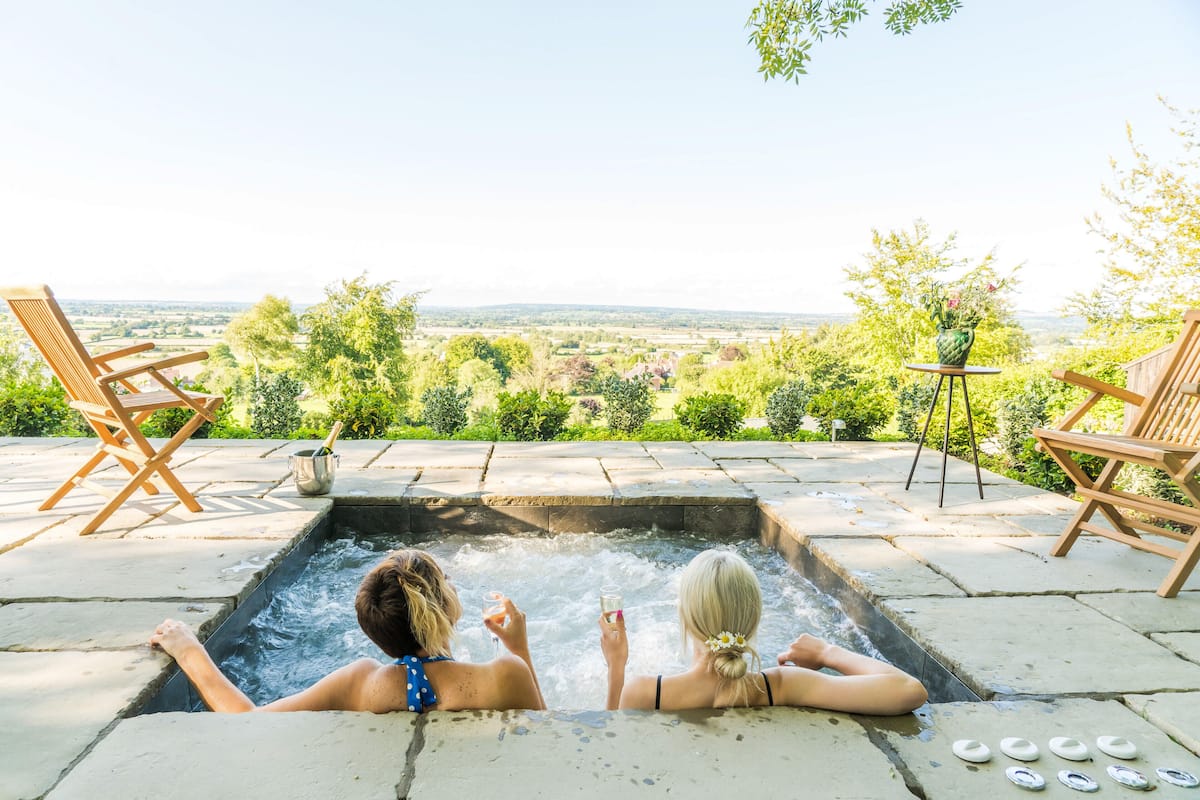 Two people relaxing in a hot tub with a scenic view of a garden and countryside.