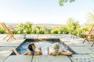 Duas pessoas relaxando em uma banheira de hidromassagem com vista panorâmica para um jardim e o campo.