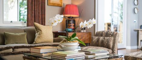 Una sala de estar con un sofá, una mesa de centro con libros y una planta, una lámpara y una silla.