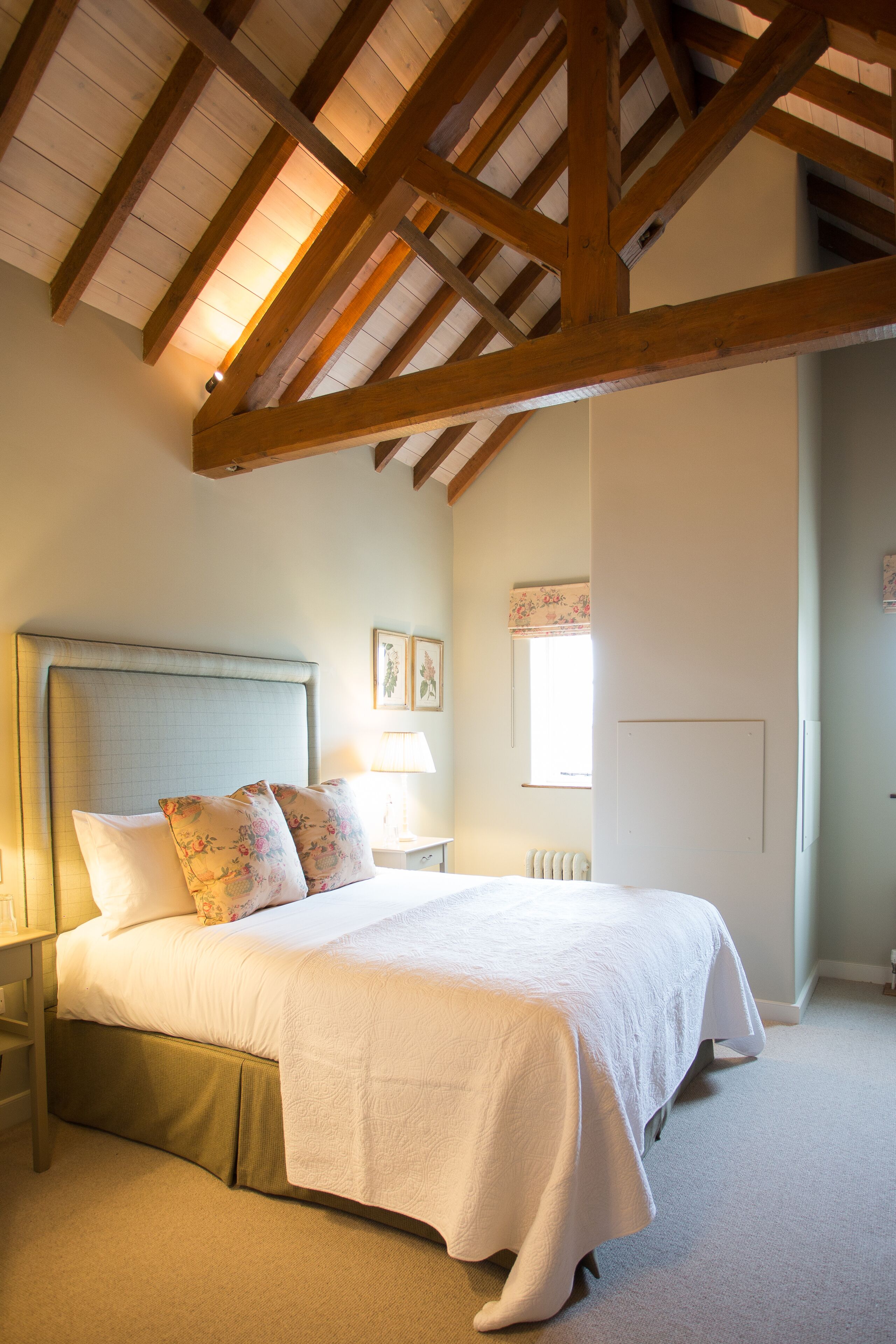 Une chambre à coucher avec un grand lit, des poutres en bois, une table de chevet et une fenêtre ornée d’un rideau à motifs floraux.