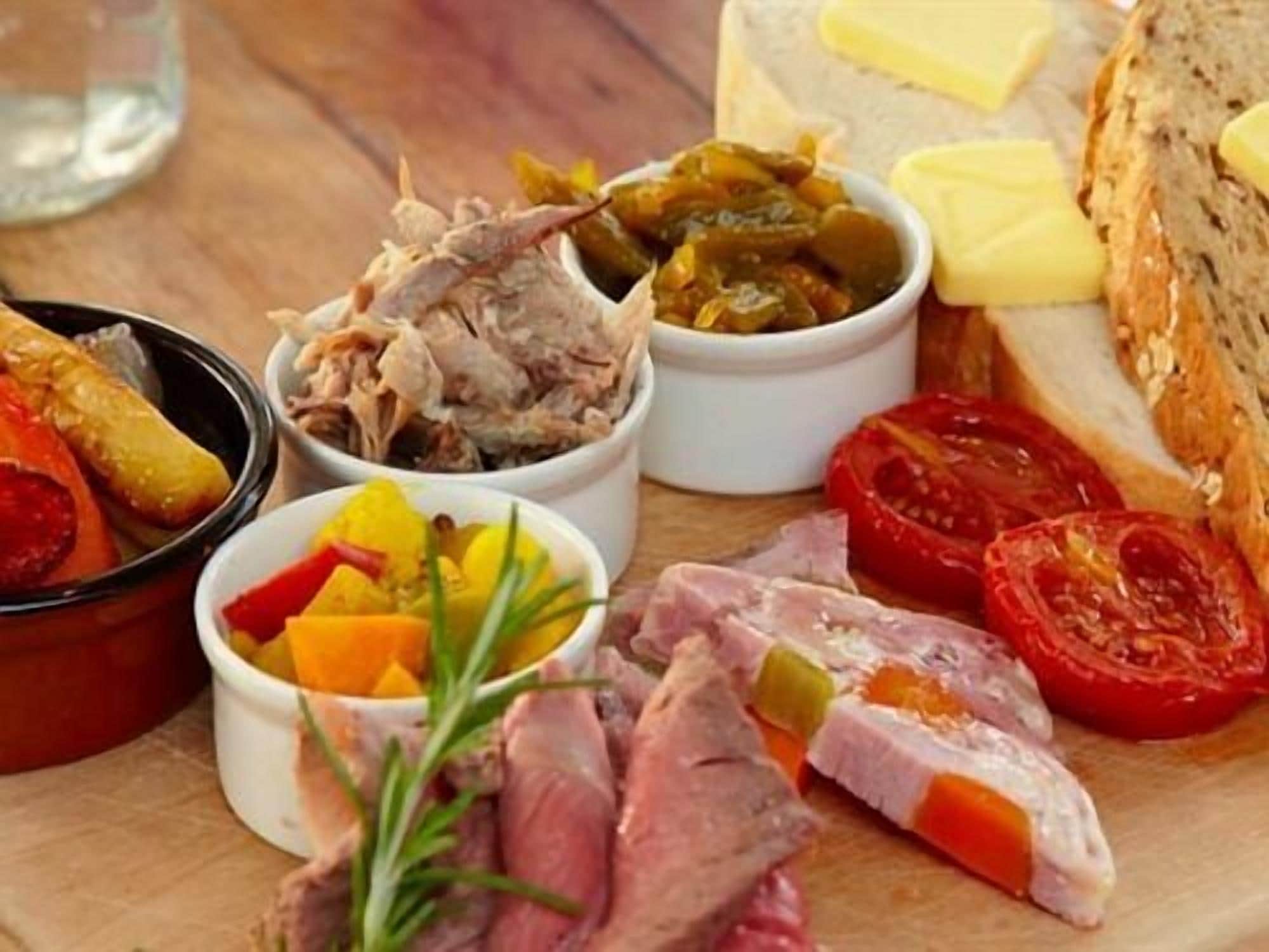 Variedad de carnes y verduras en una tabla de madera, incluyendo tomates en rodajas, pepinillos y un recipiente que parece ser un guiso de carne.