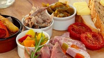 Variedad de carnes y verduras en una tabla de madera, incluyendo tomates en rodajas, pepinillos y un recipiente que parece ser un guiso de carne.