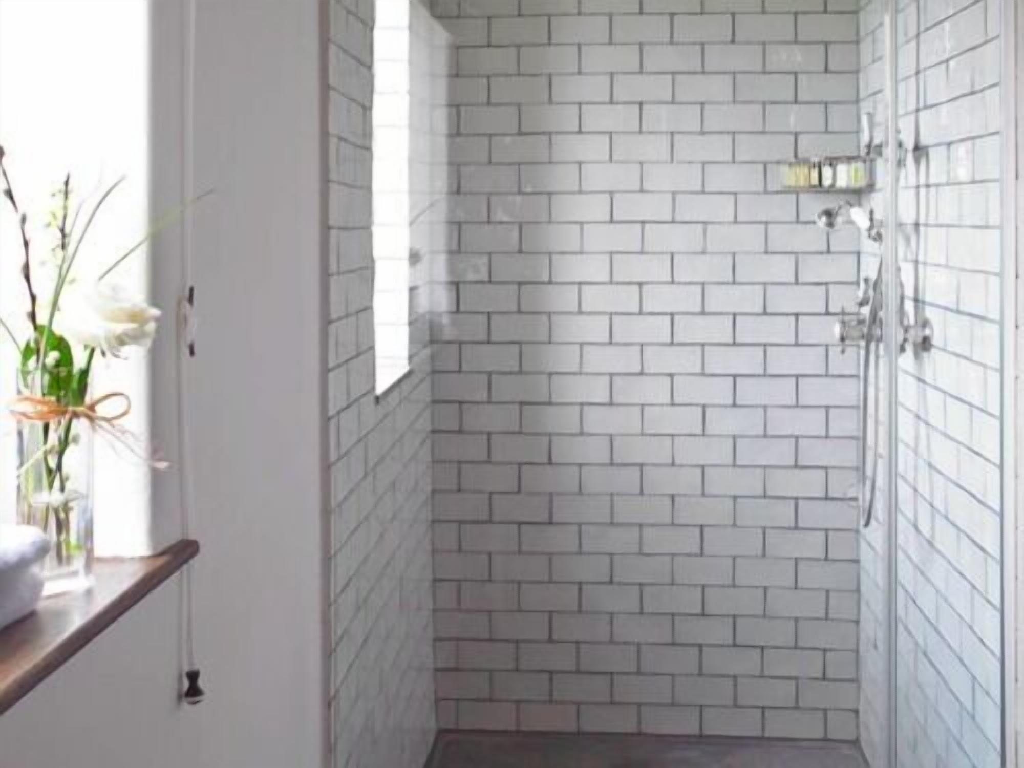 A small, enclosed shower area with white tiled walls and a wooden shelf with a vase of flowers.