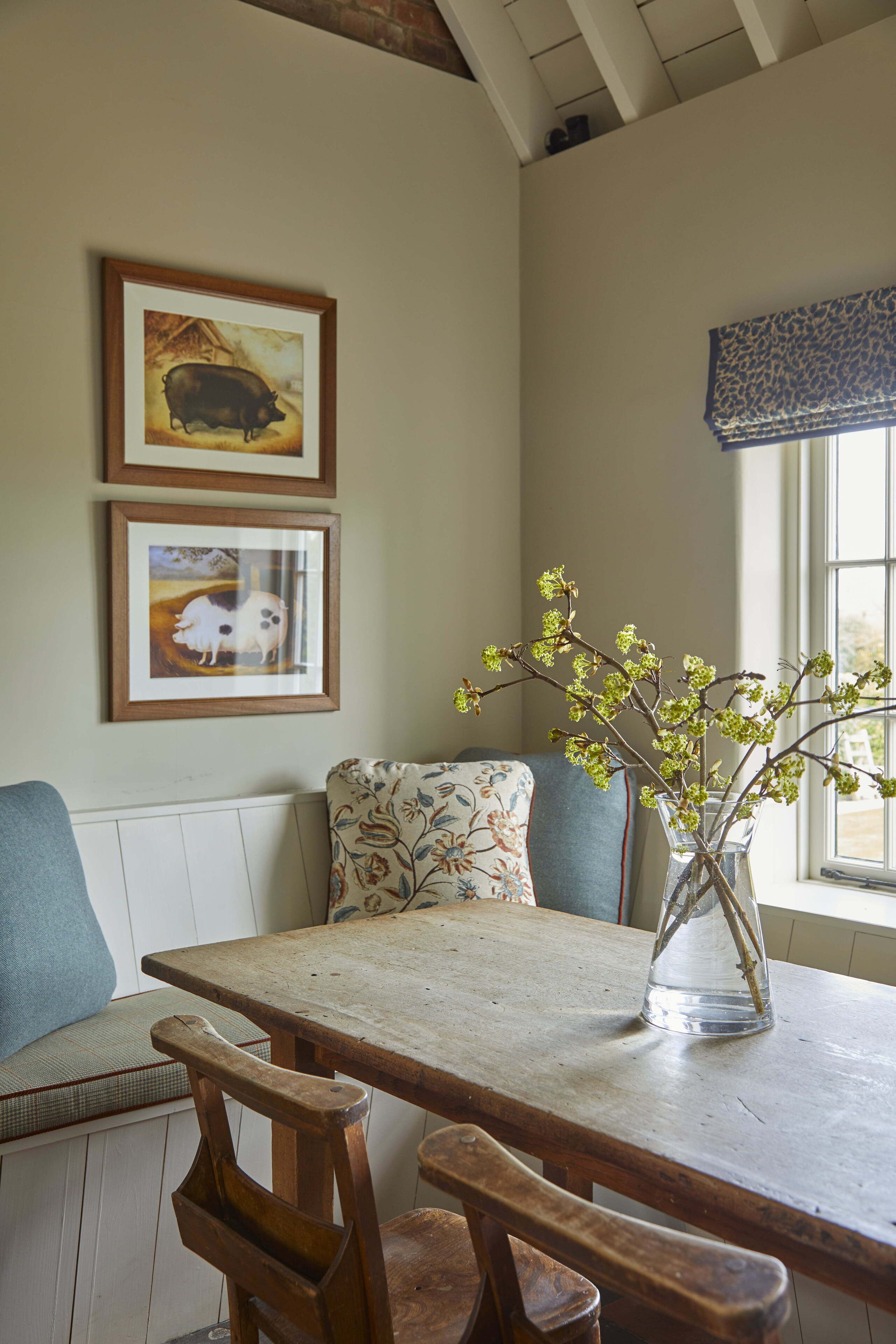 A dining area with a wooden table, two framed animal paintings, a vase with branches, and a window with a blue valance.