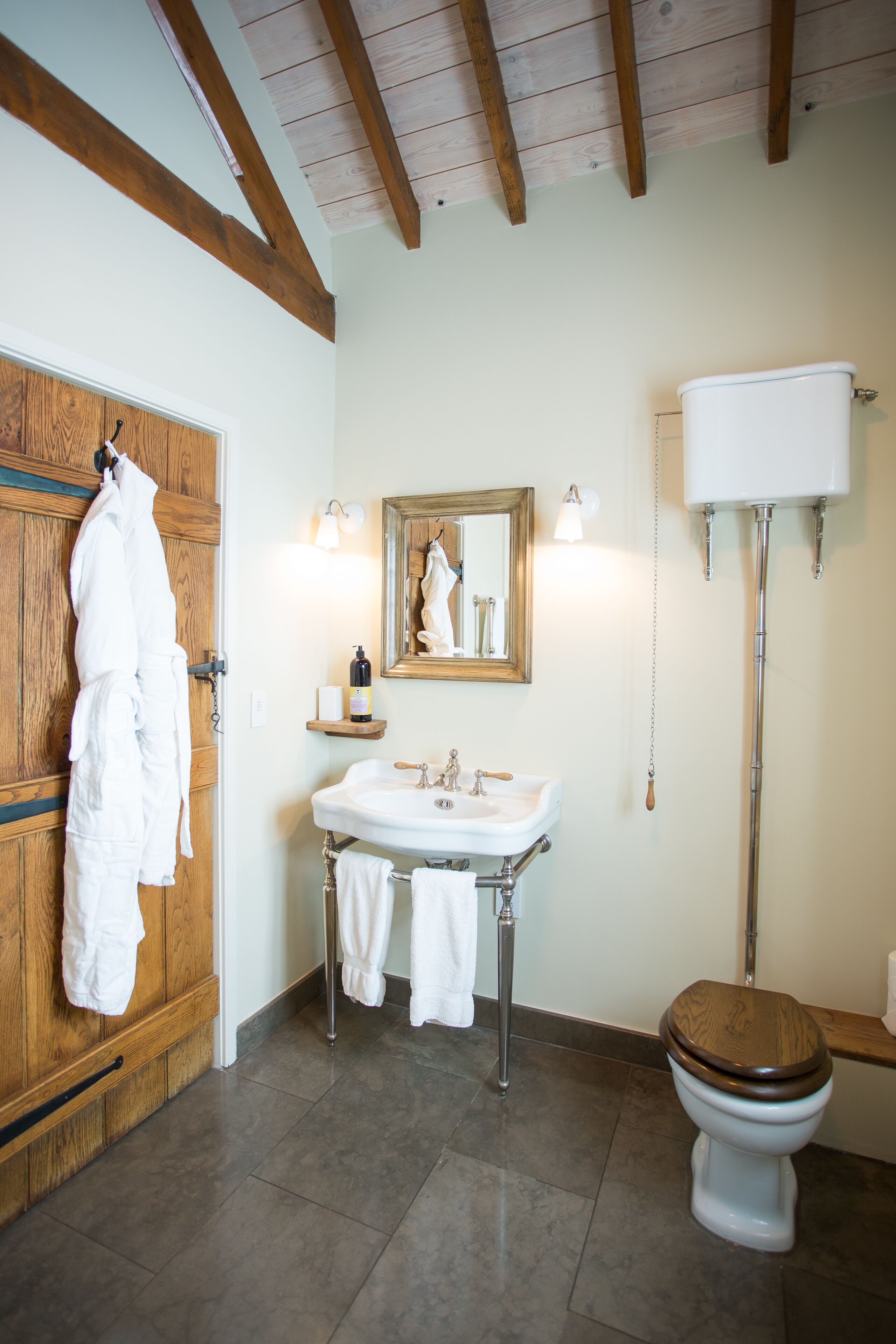 Ein Badezimmer mit Holztür, weißem Waschbecken mit Spiegel, Toilette und einer Badmatte aus Holz.
