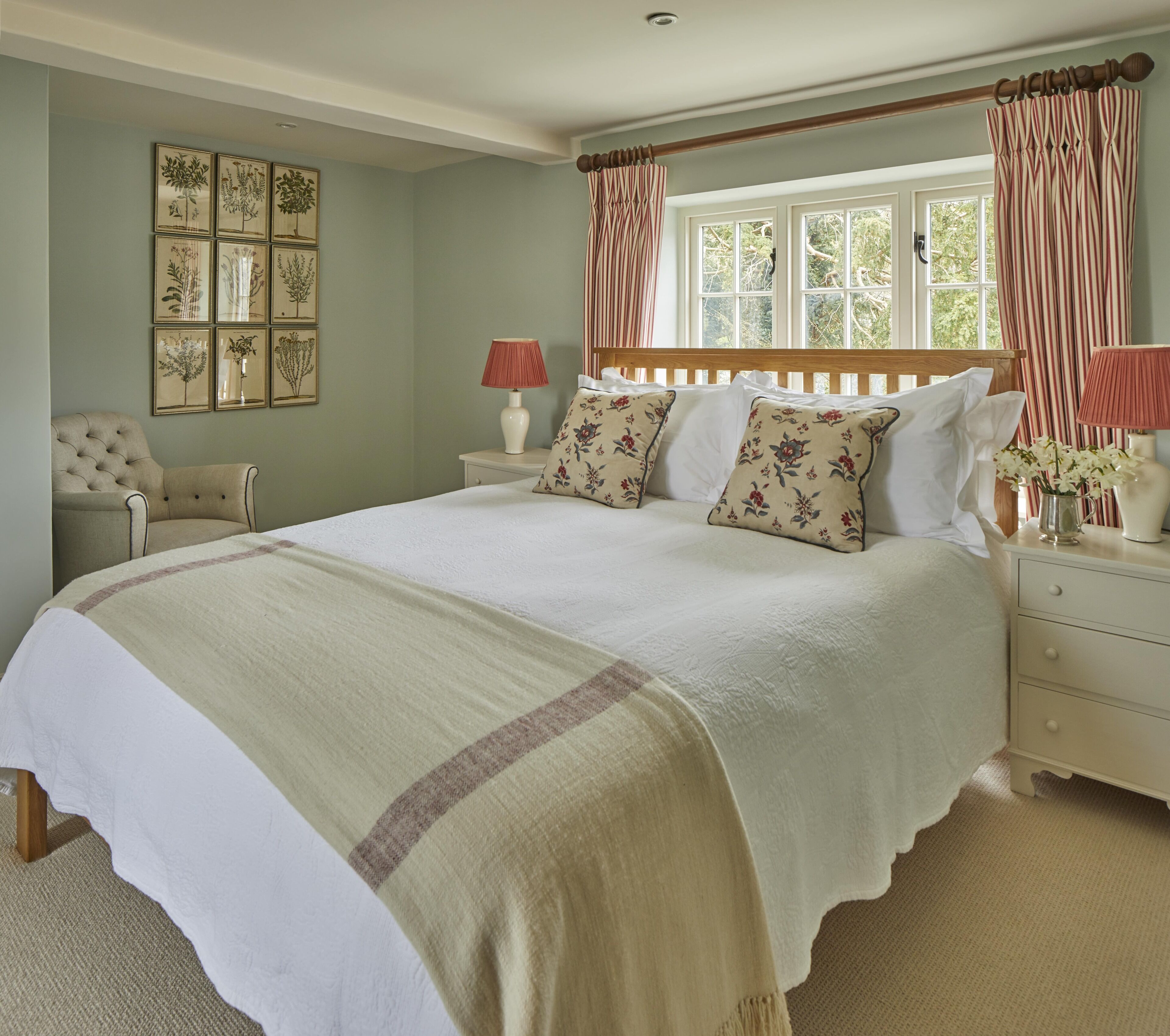 A bedroom with a large bed, two bedside tables, a chair, a window with curtains, and framed botanical prints on the wall.
