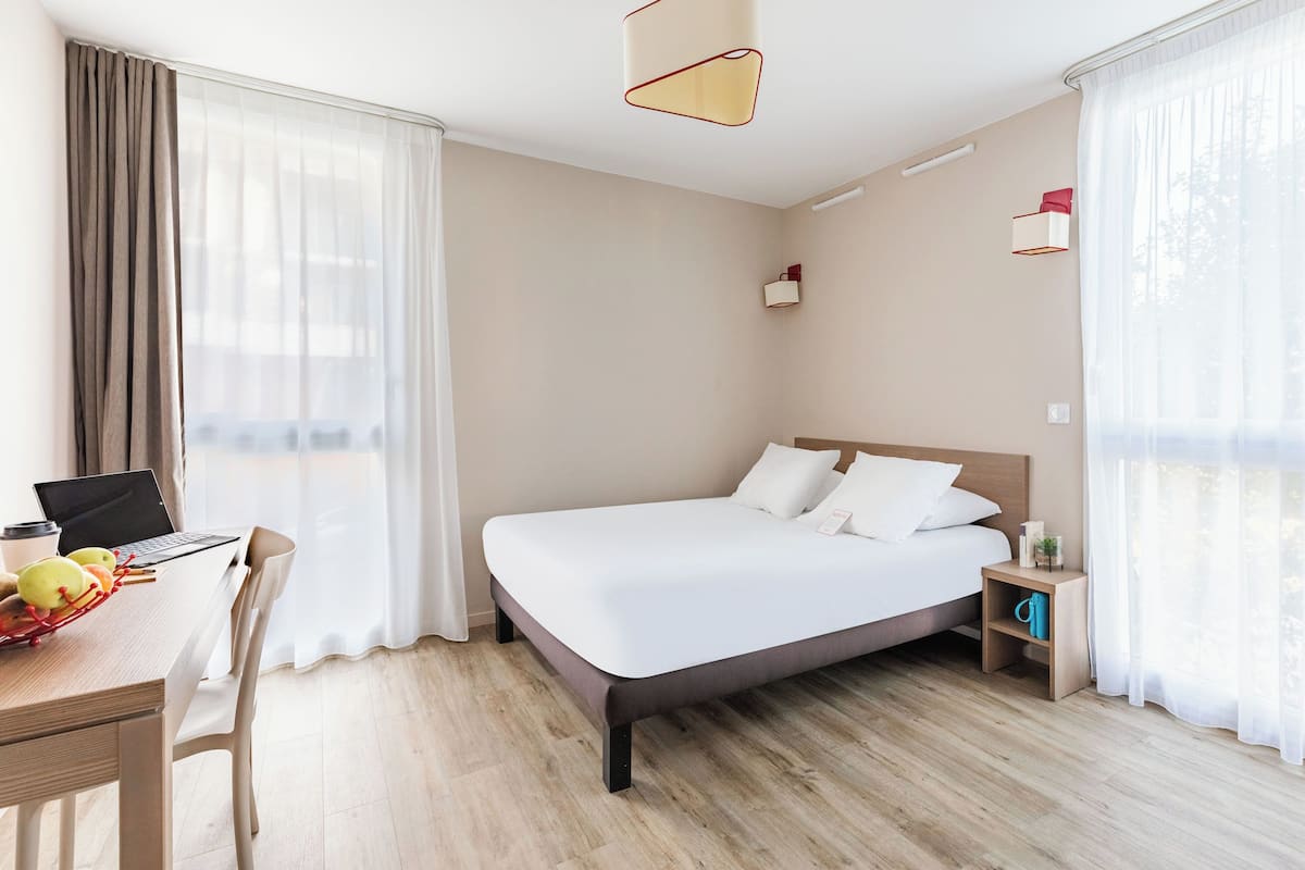 Ein Hotelzimmer mit einem Bett, einem Schreibtisch mit einem Laptop, einem Sessel, einem kleinen Tisch mit Früchten und einem Nachttisch mit einer Lampe.