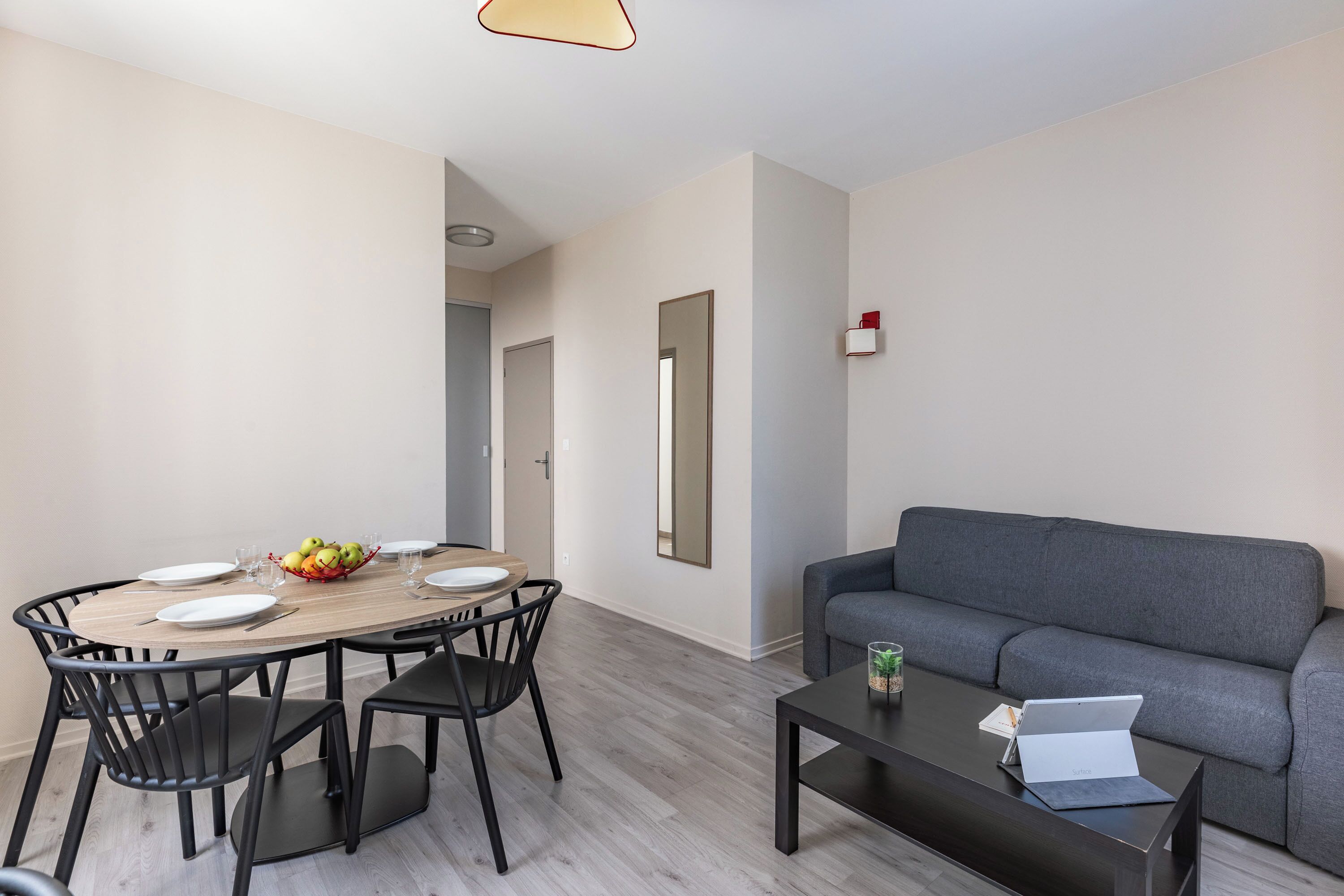 Una sala de estar moderna con un sofá gris, una mesa de comedor de madera con sillas y una mesa baja con una computadora portátil.