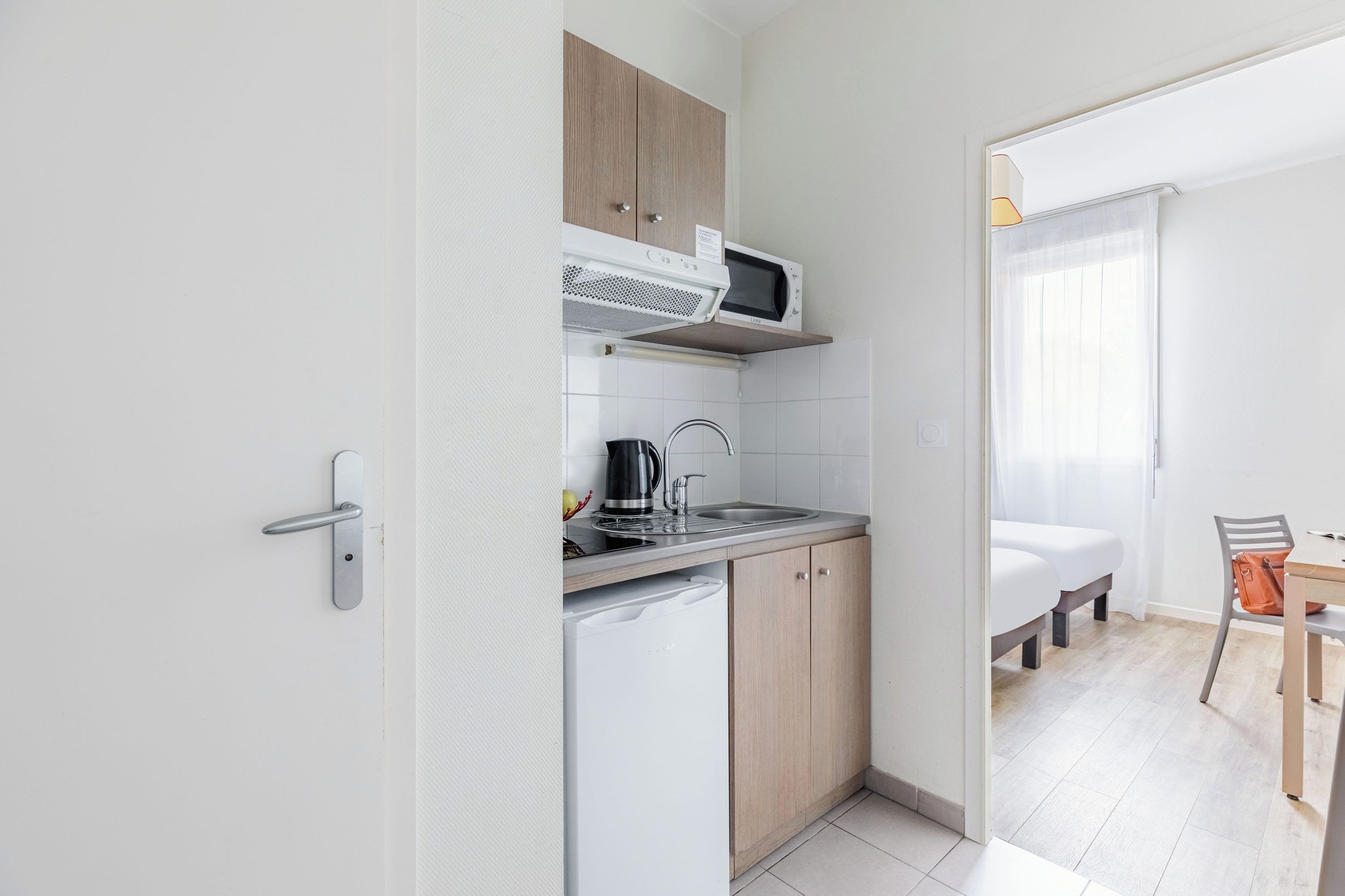 A compact kitchenette with a microwave, sink, and a small refrigerator. There is a wooden cabinet above the microwave and a chair with a bag next to a table.