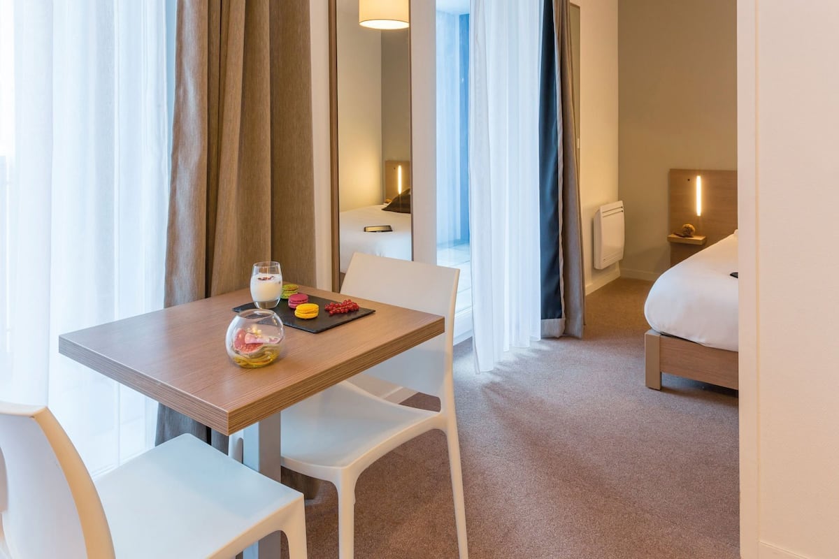 Ein Hotelzimmer mit Esstisch, Stühlen, einem Bett und einem kleinen Nachttisch mit Lampe.