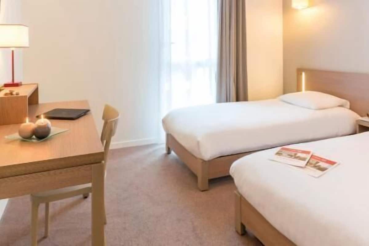 Ein Hotelzimmer mit Schreibtisch, zwei Betten, einem Sessel, einer Lampe und einem fenster mit Vorhängen.