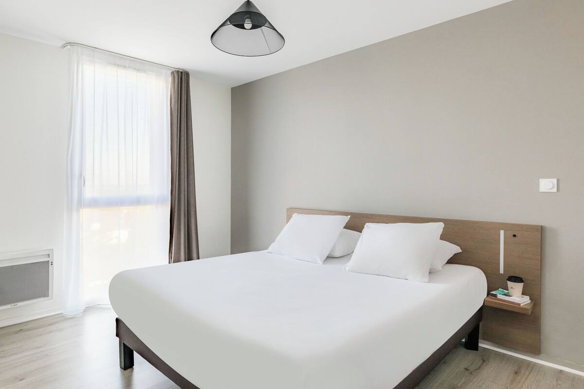 Ein modernes Hotelzimmer mit einem großen Bett, einem Nachttisch mit Lampe und Blick ins Freie durch transparente Vorhänge.