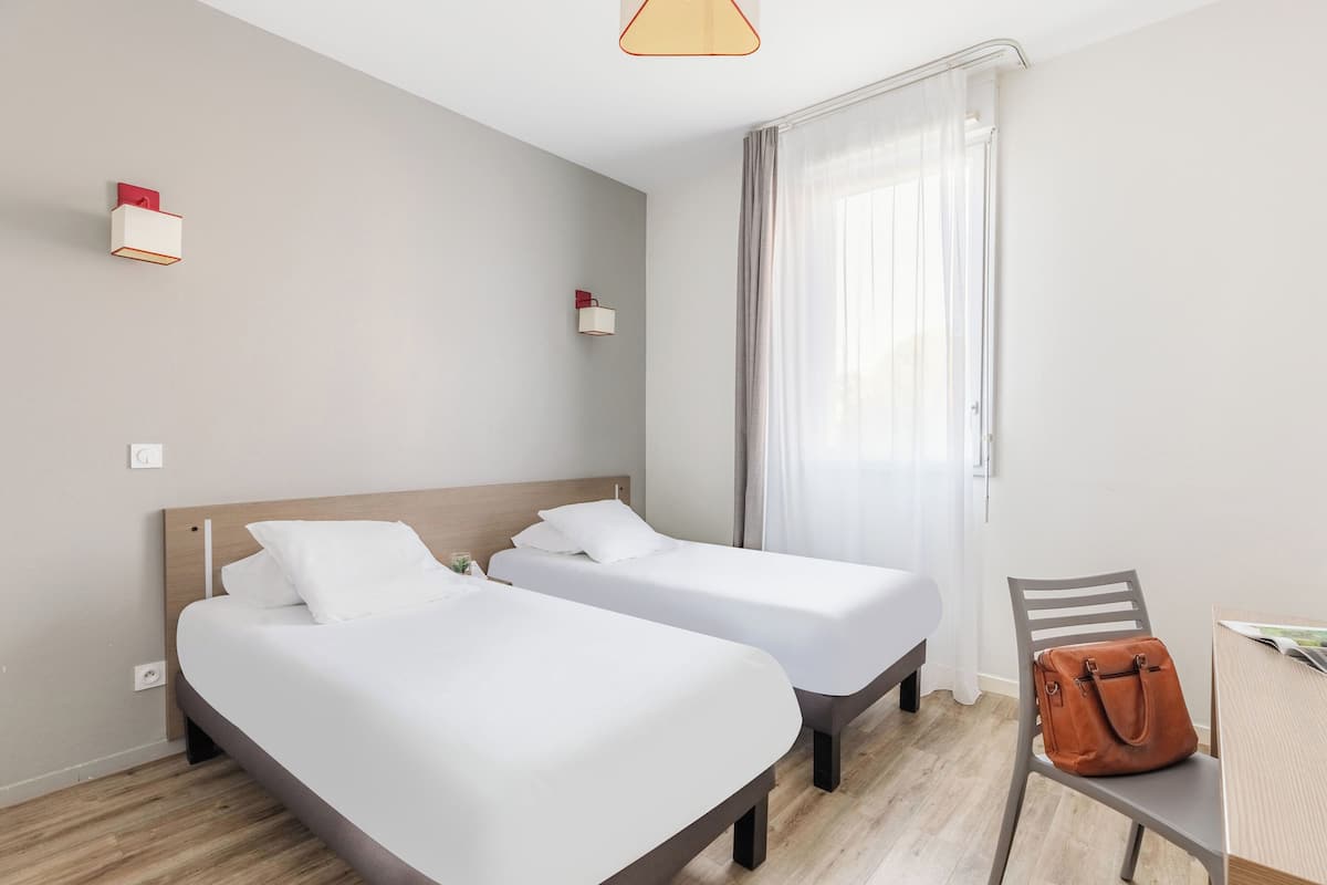 Ein Hotelzimmer mit zwei Betten, Holzboden, einem Tisch mit Stuhl und einem Fenster mit Vorhängen.