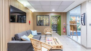 Un espace d’accueil moderne avec un canapé gris, des fauteuils en osier, une table basse en bois et un téléviseur à écran plat fixé au mur.
