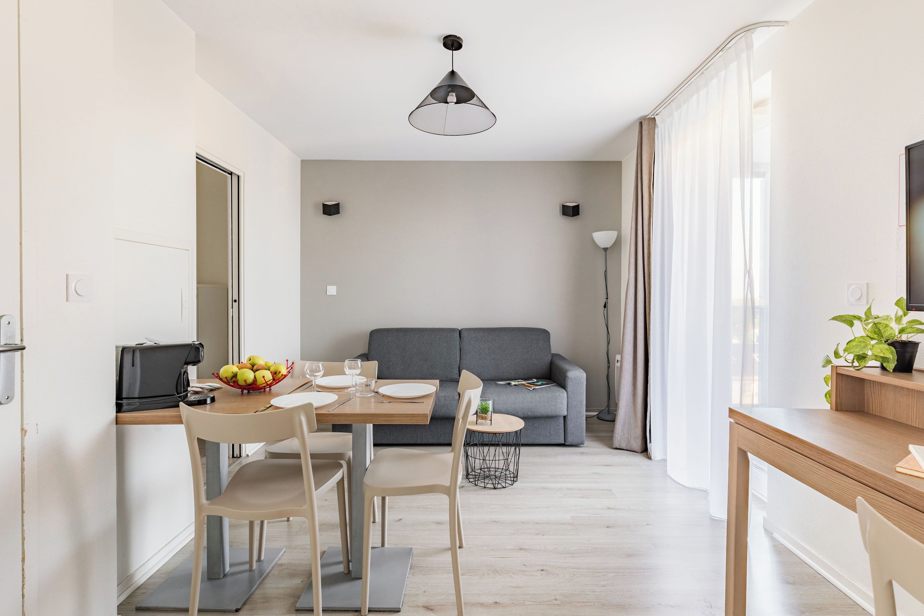 Een moderne keuken met een eettafel, stoelen en een schaal fruit. Er is een grijze bank en een klein bijzettafeltje met een plantje op de achtergrond.