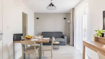 Een moderne keuken met een eettafel, stoelen en een schaal fruit. Er is een grijze bank en een klein bijzettafeltje met een plantje op de achtergrond.