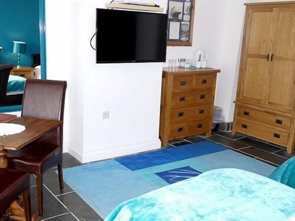 A bedroom with a bed, a wooden dresser, a television mounted on the wall, and a blue rug. - Sawyers Bed and Breakfast (Looe)