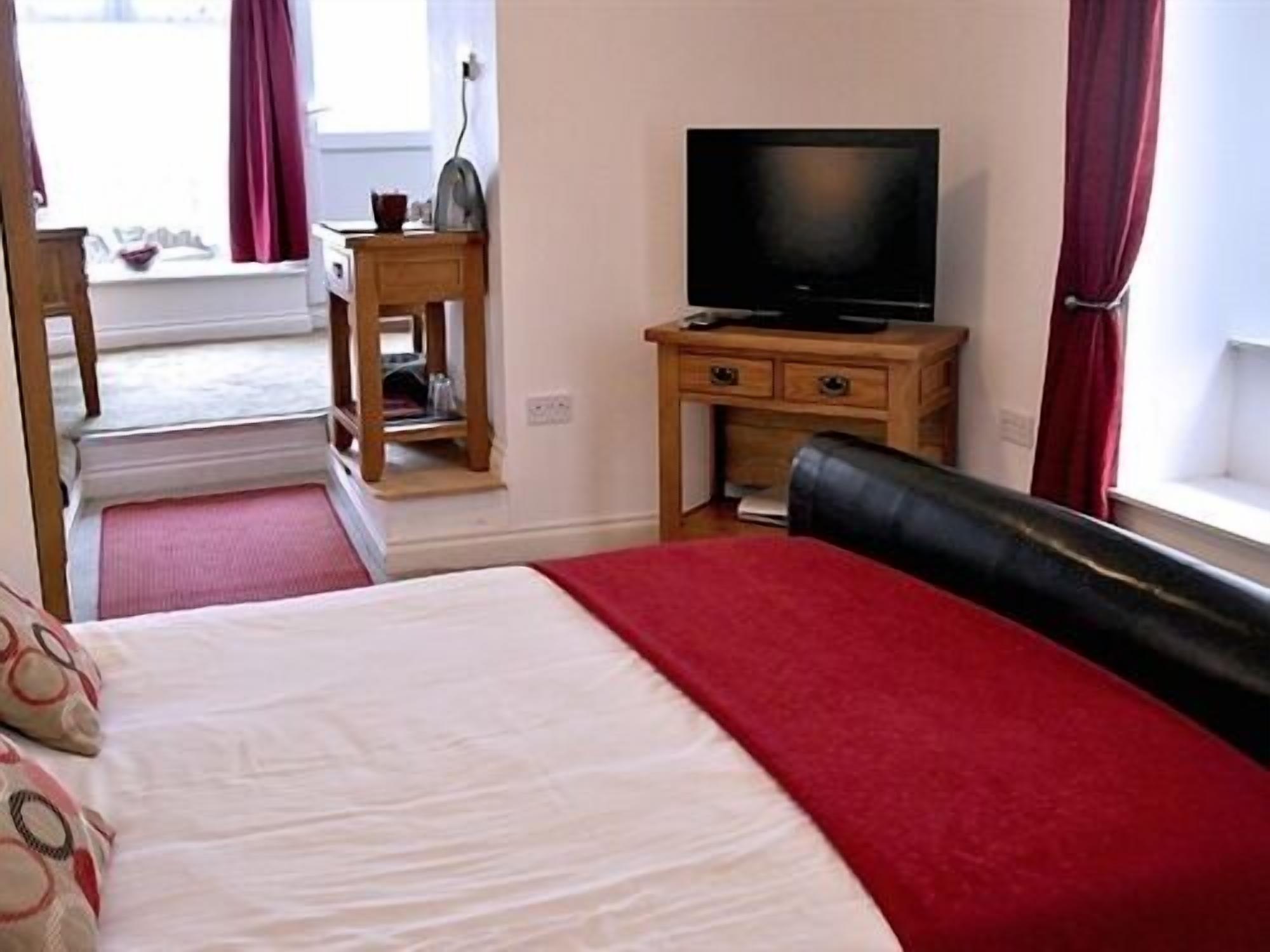 A hotel room with a bed, a television on a wooden stand, a small table with a lamp, and a window with curtains.