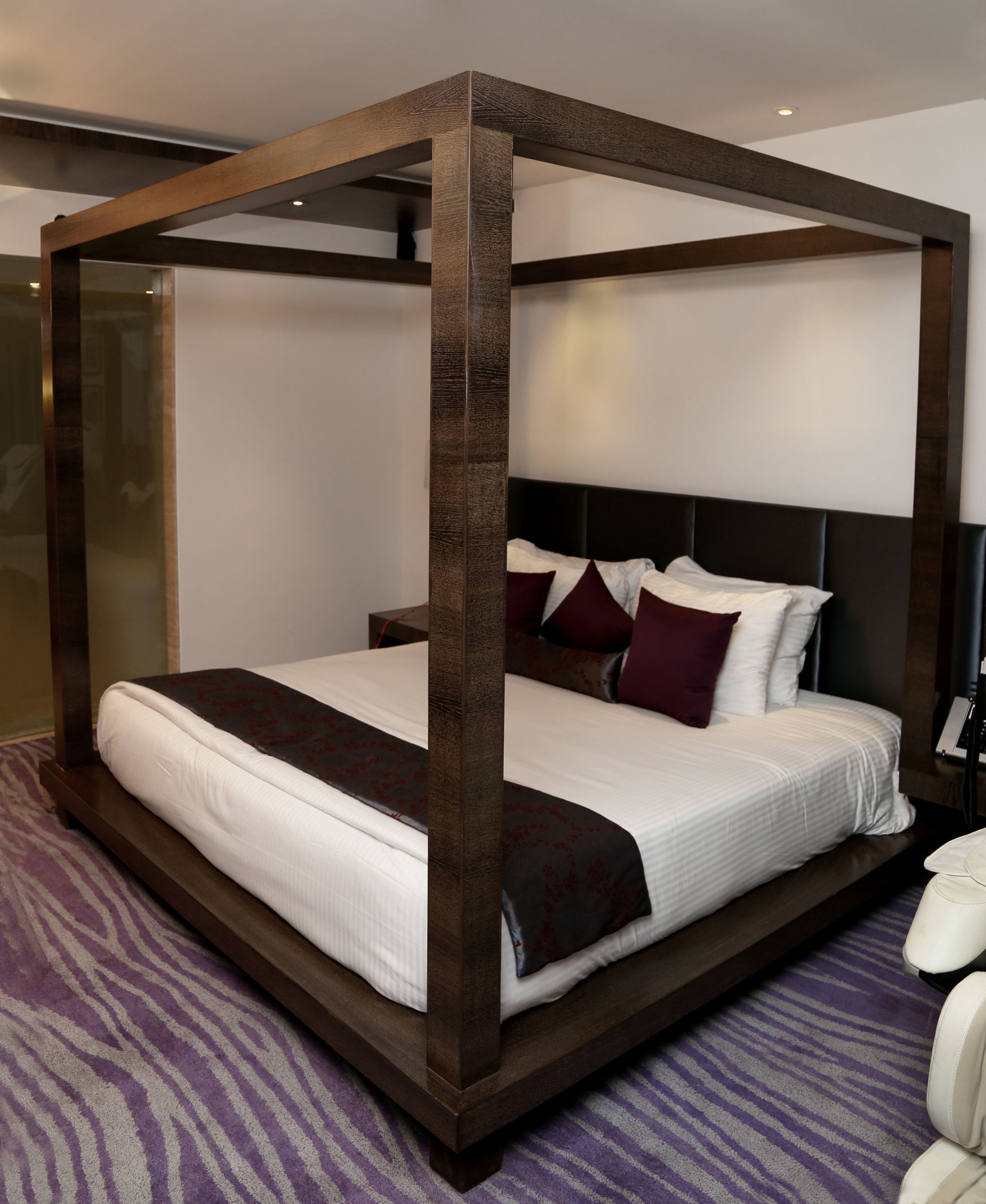 Una cama con dosel de madera oscura, ropa de cama blanca y una alfombra a rayas moradas.