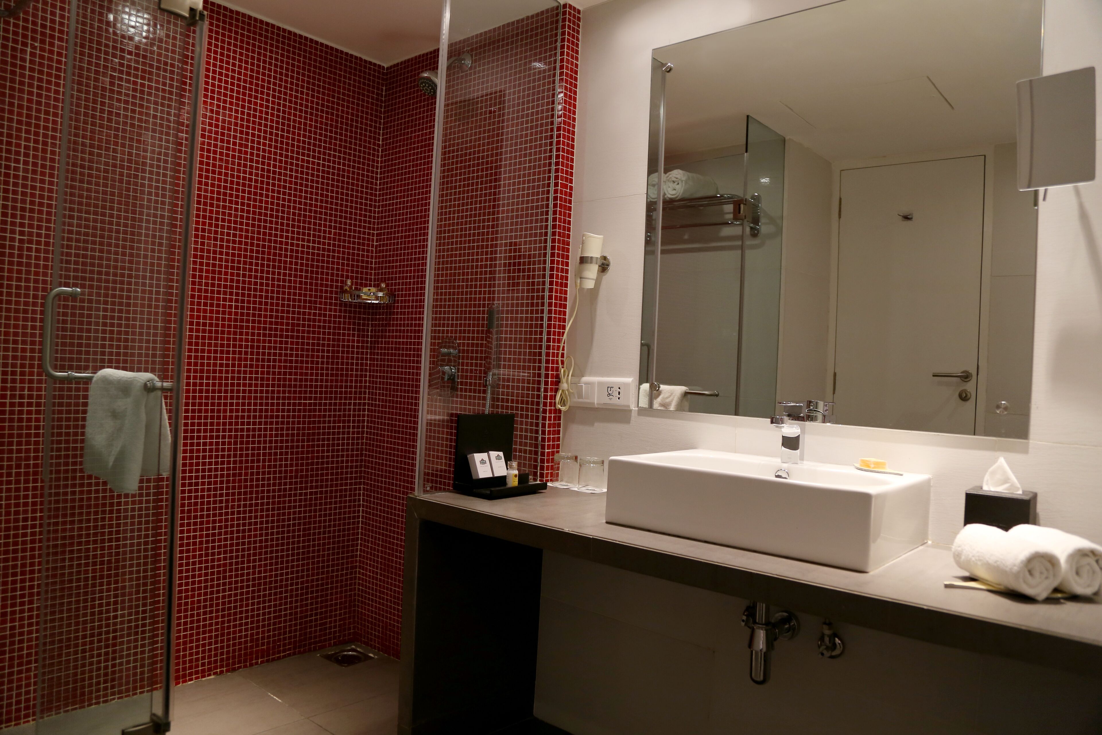 Un baño moderno con pared de ducha de azulejos rojos, un lavabo blanco con espejo arriba y un perchero con toallas.
