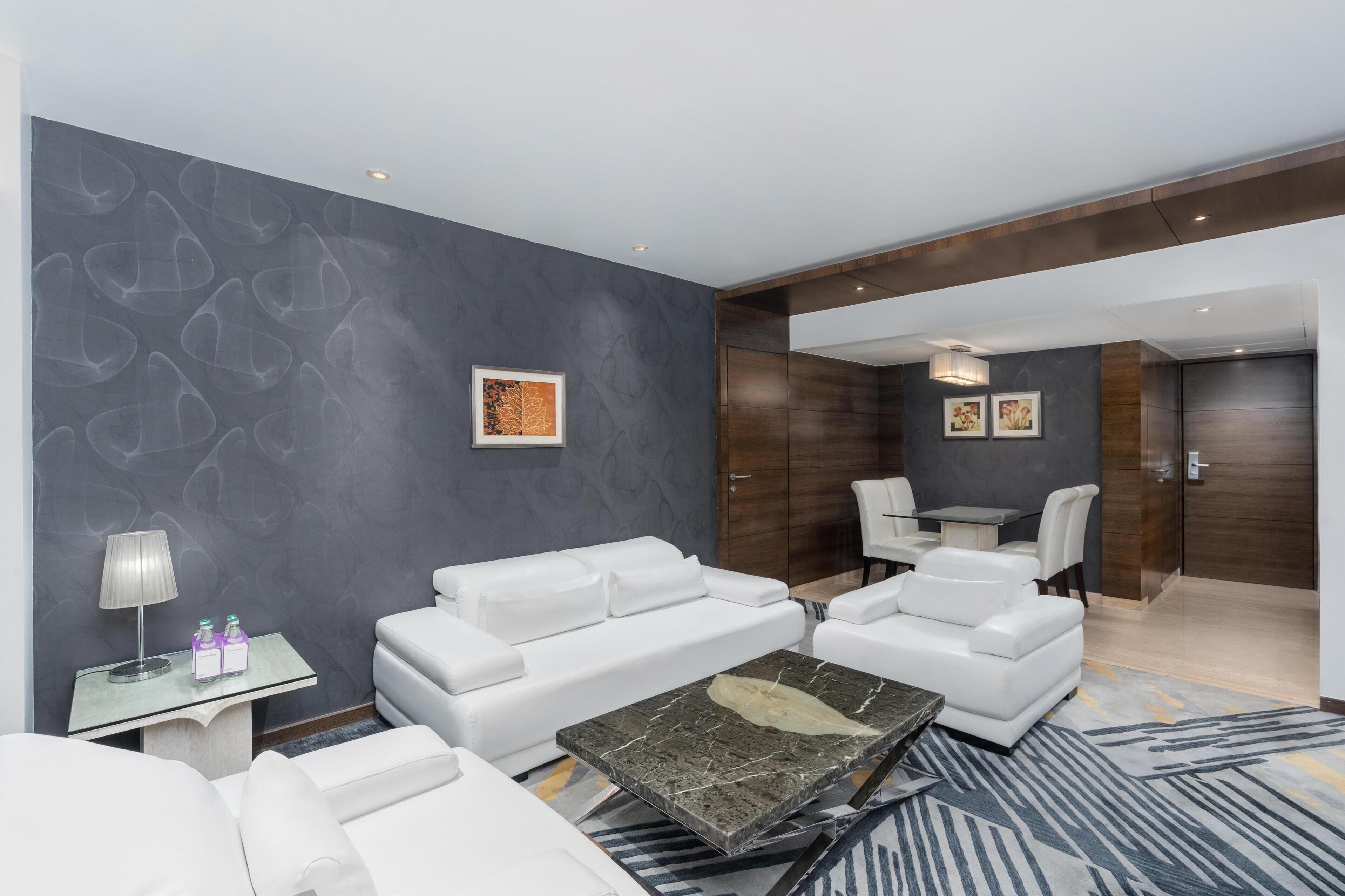A modern hotel room with a grey textured wall, white leather sofa, a marble coffee table, and a small side table with a lamp and bottles.