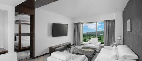 A modern hotel room with a large window, a flat-screen TV, a sofa, a coffee table, and a bed with a headboard.