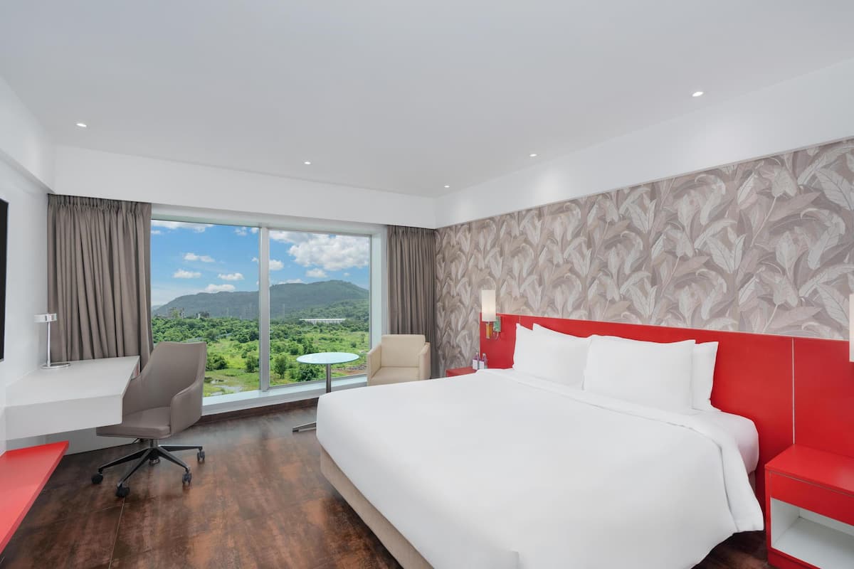 A hotel room with a large bed, a desk, a chair, and a view of greenery and mountains through the window.