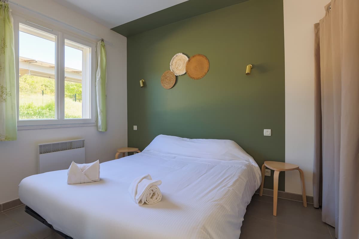 A neatly made bed with white linens, a small wooden stool, and a wall with three woven placemats.