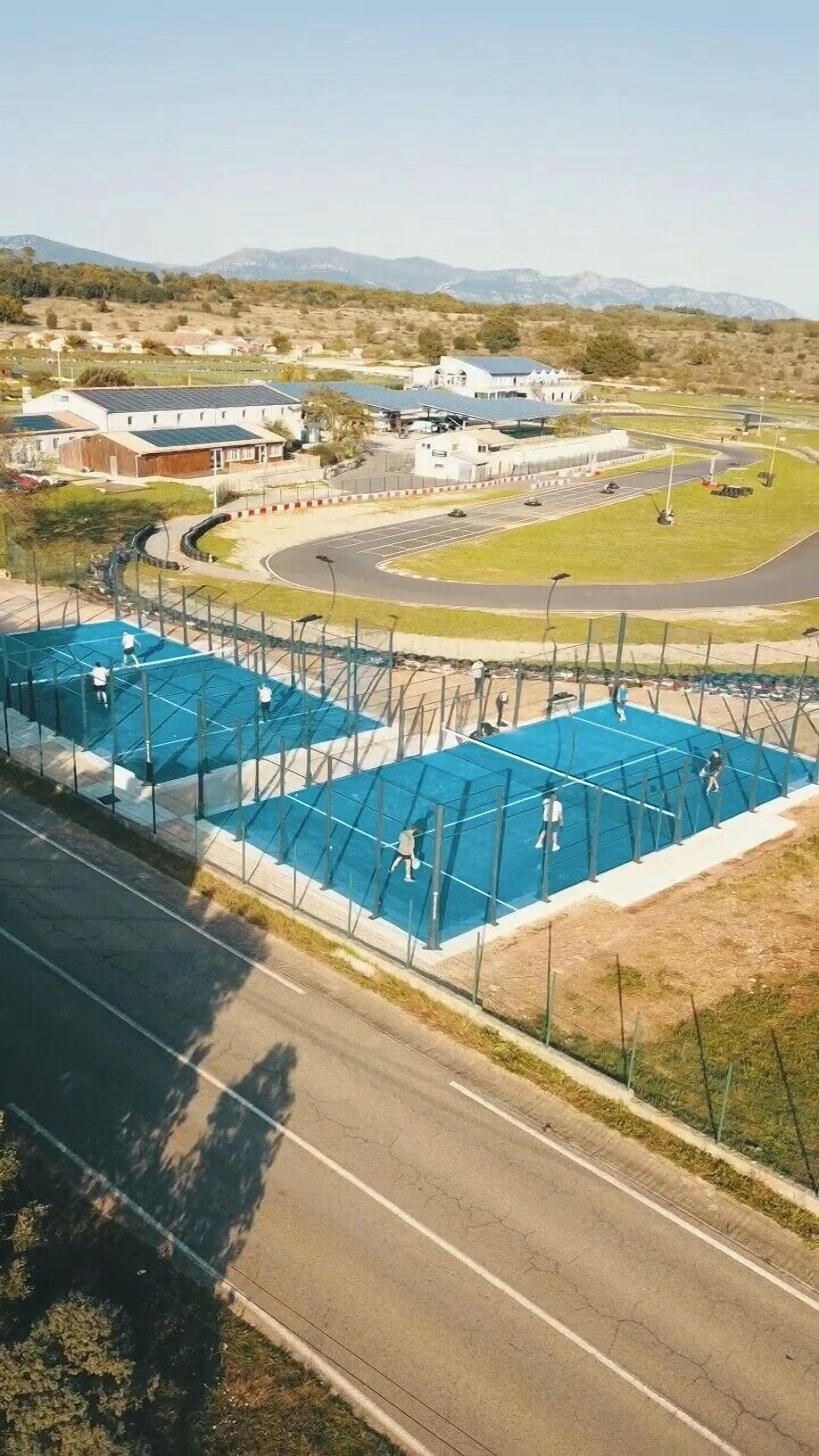 An aerial view of a sports facility with multiple tennis courts, a parking area, and a building with solar panels.
