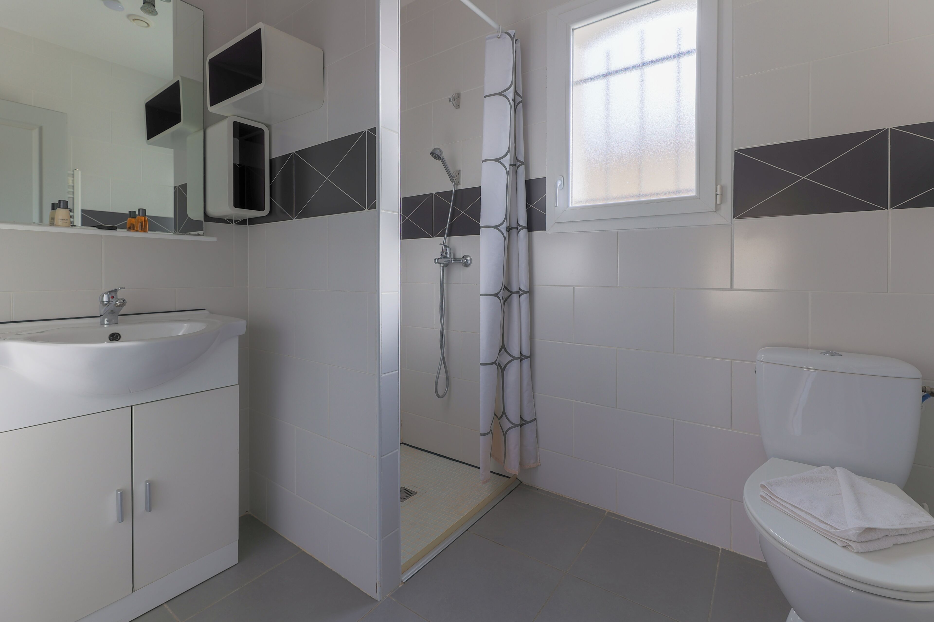 Un bany amb un lavabo blanc, un vàter, una dutxa amb porta de vidre i un mirall amb una balda a la part superior.