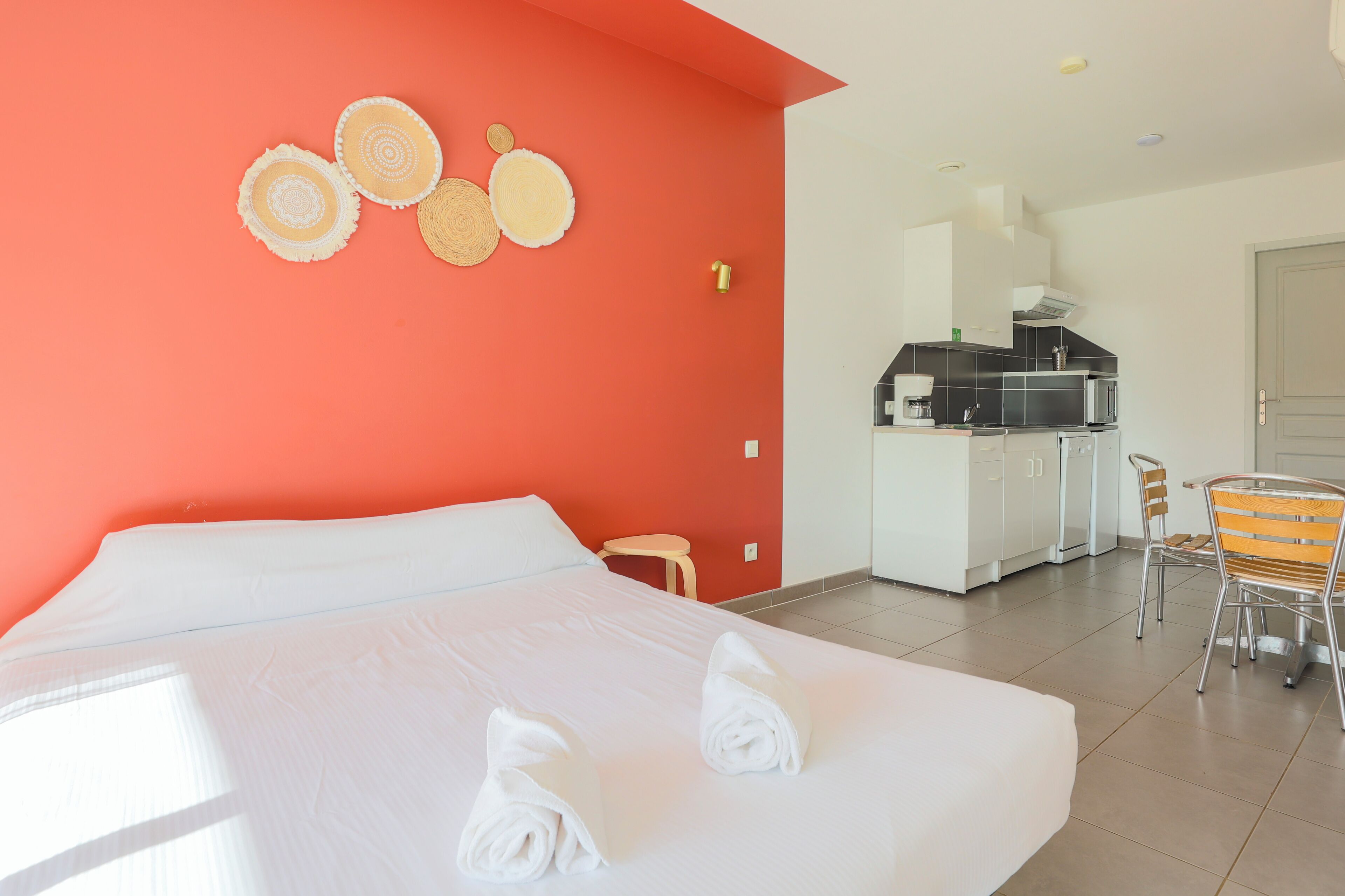 Une chambre d’hôtel comprenant un lit, un petit tabouret, une table à manger avec des chaises et un coin cuisine équipé d’un four à micro-ondes et d’un grille-pain.