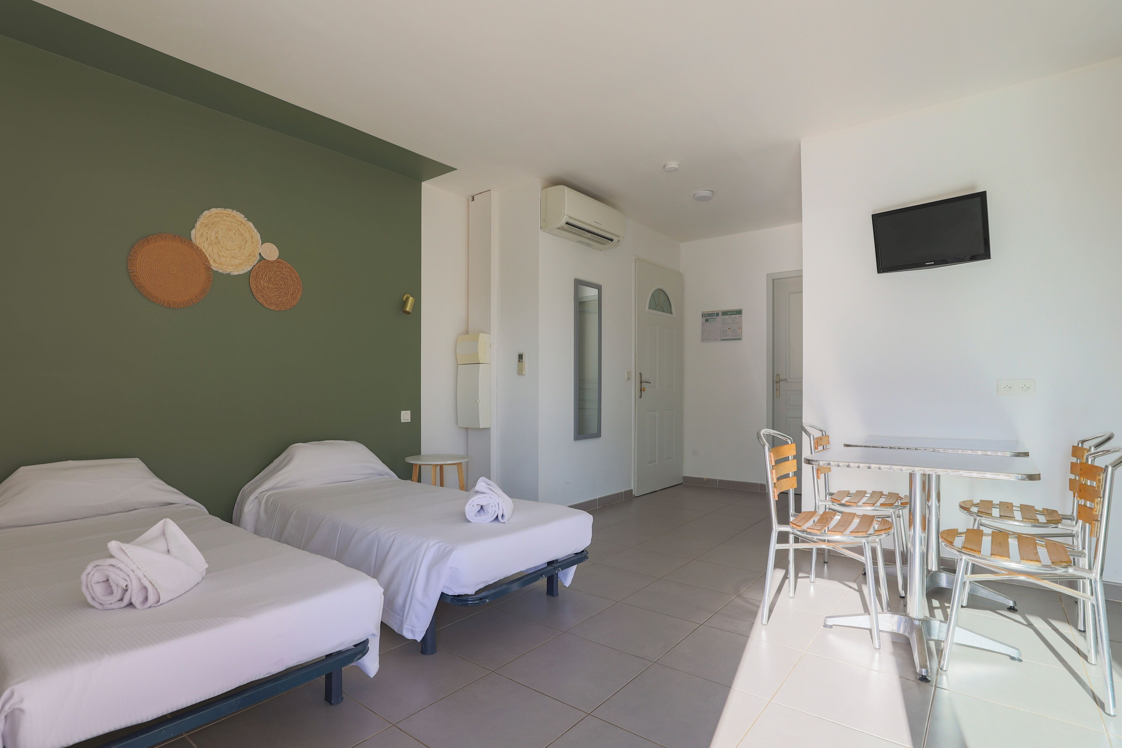 A hotel room with two beds, a small table with chairs, a TV, and a wall-mounted air conditioner.