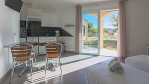 A modern hotel room with a kitchen area, a dining table with chairs, a bed, and a large sliding glass door leading to a balcony with a view of the outdoors. - Résidence du Parc - Gorges de l'Hérault - Cévennes - Parc Sports & Loisirs Brissac Ganges (Brissac)